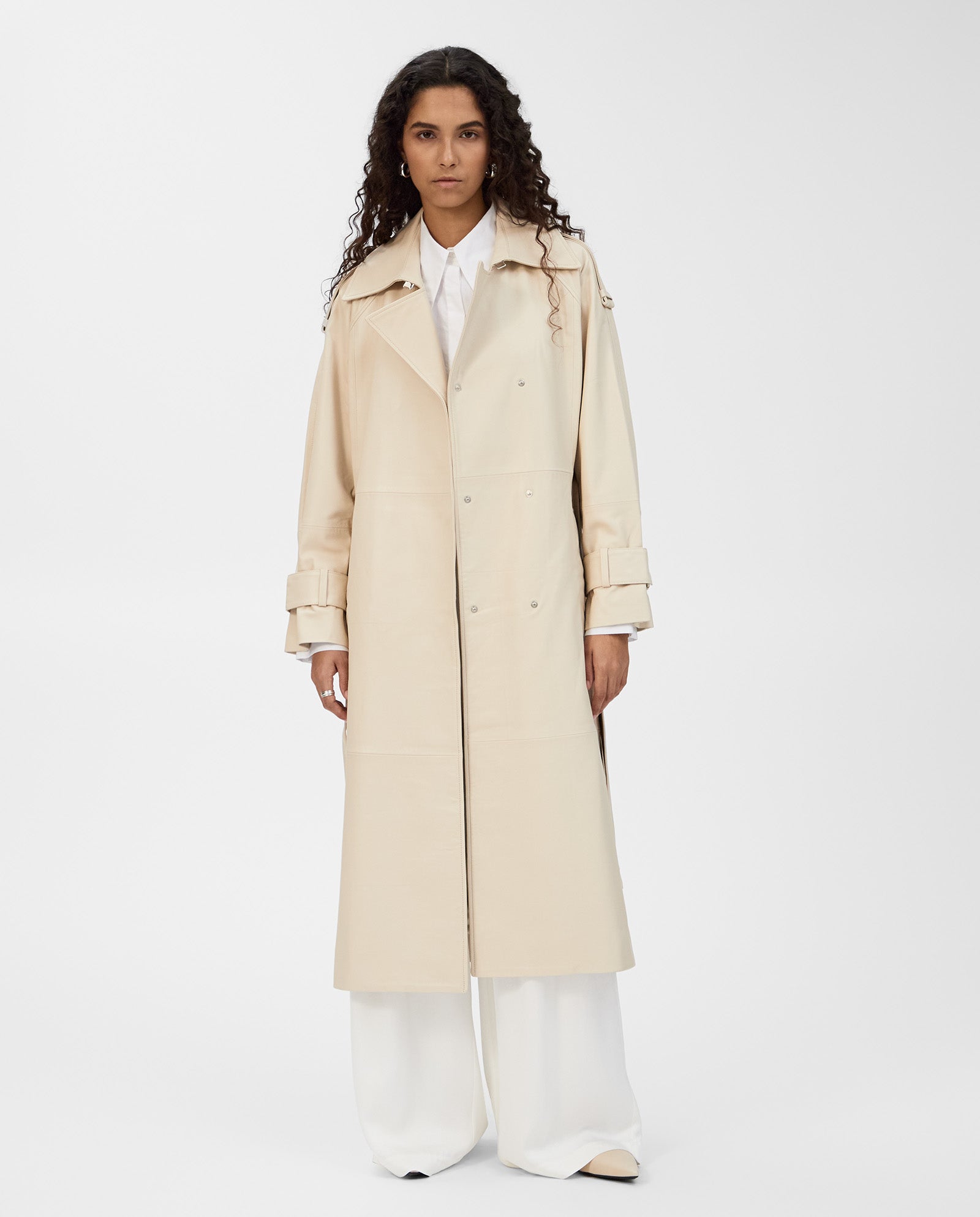 Wearing the IVY OAK LILITH Coat, a classic long beige trench with storm flap, plus a white shirt and wide-leg white pants, a person stands against a plain background.