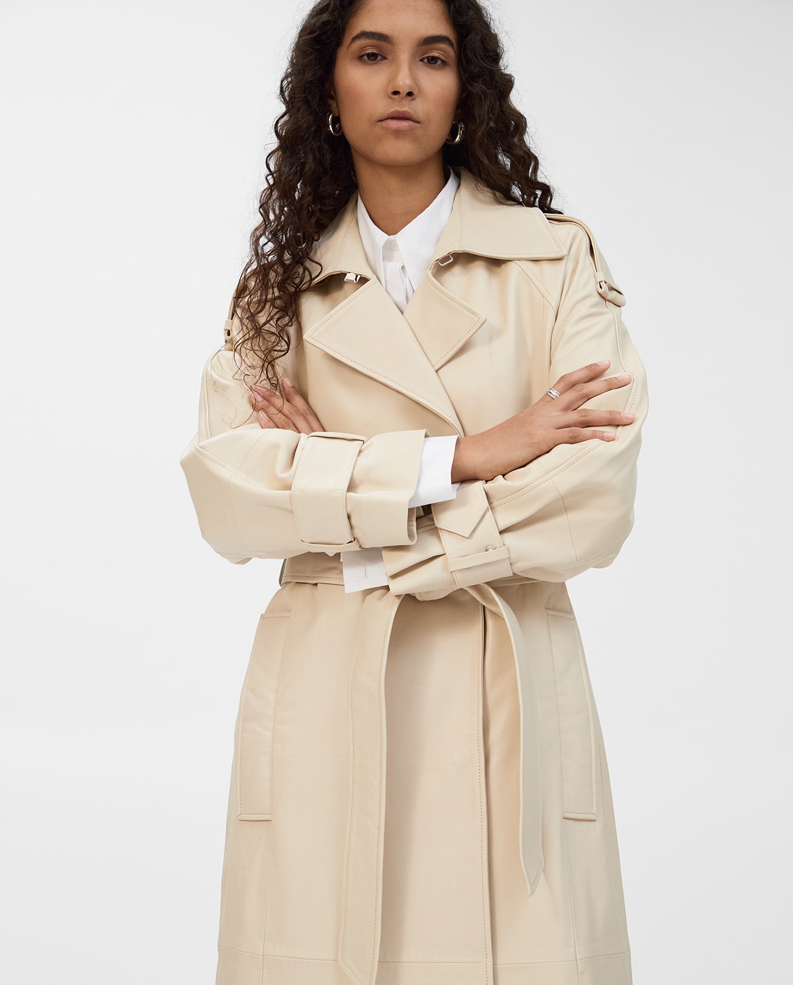 A woman with long curly hair wears the IVY OAK LILITH Coat, a classic beige trench with a storm flap, over a white shirt. She stands facing forward with arms crossed.