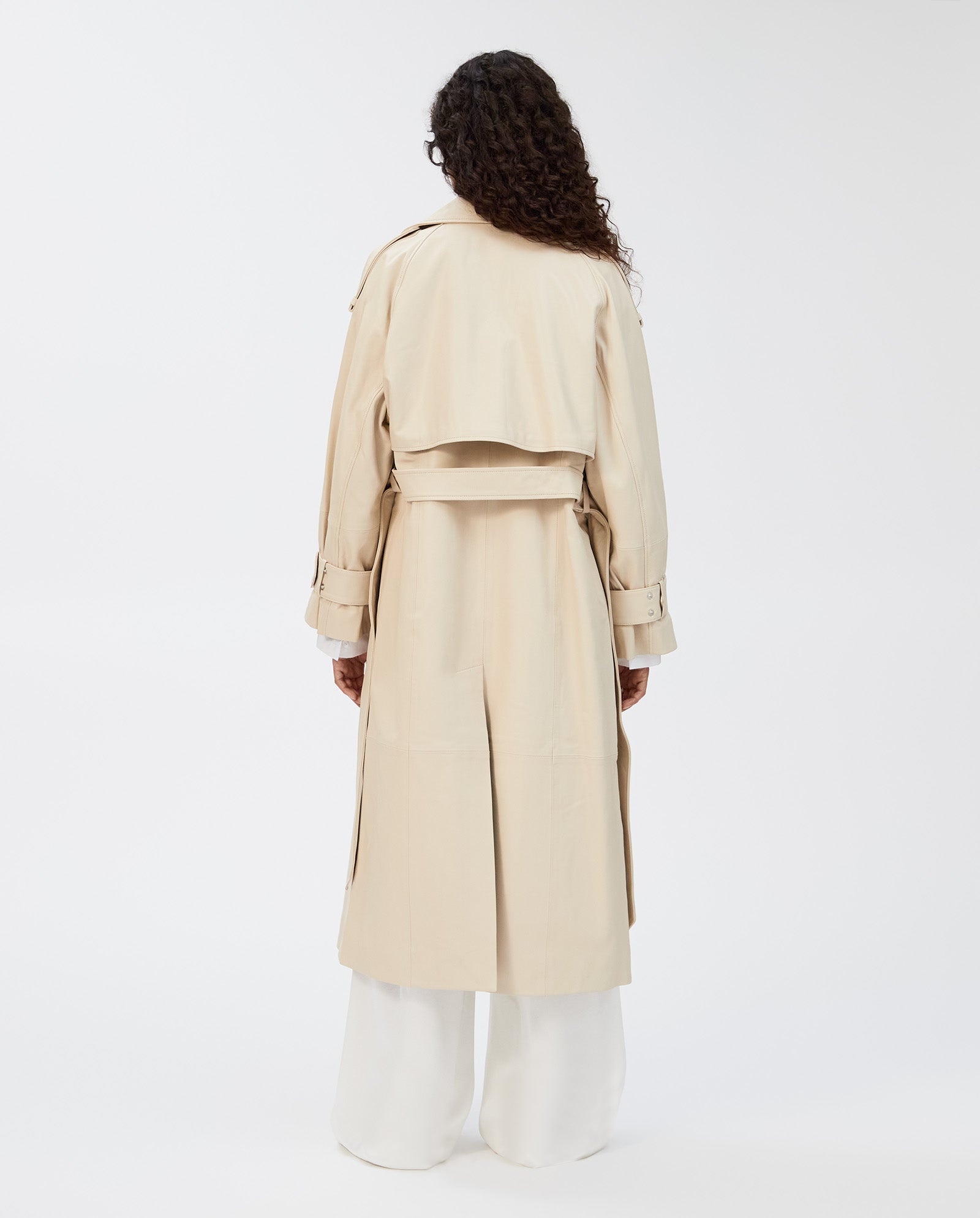 A person with long curly hair is seen from behind wearing the IVY OAK LILITH Coat in classic beige, featuring a storm flap, paired with white pants.