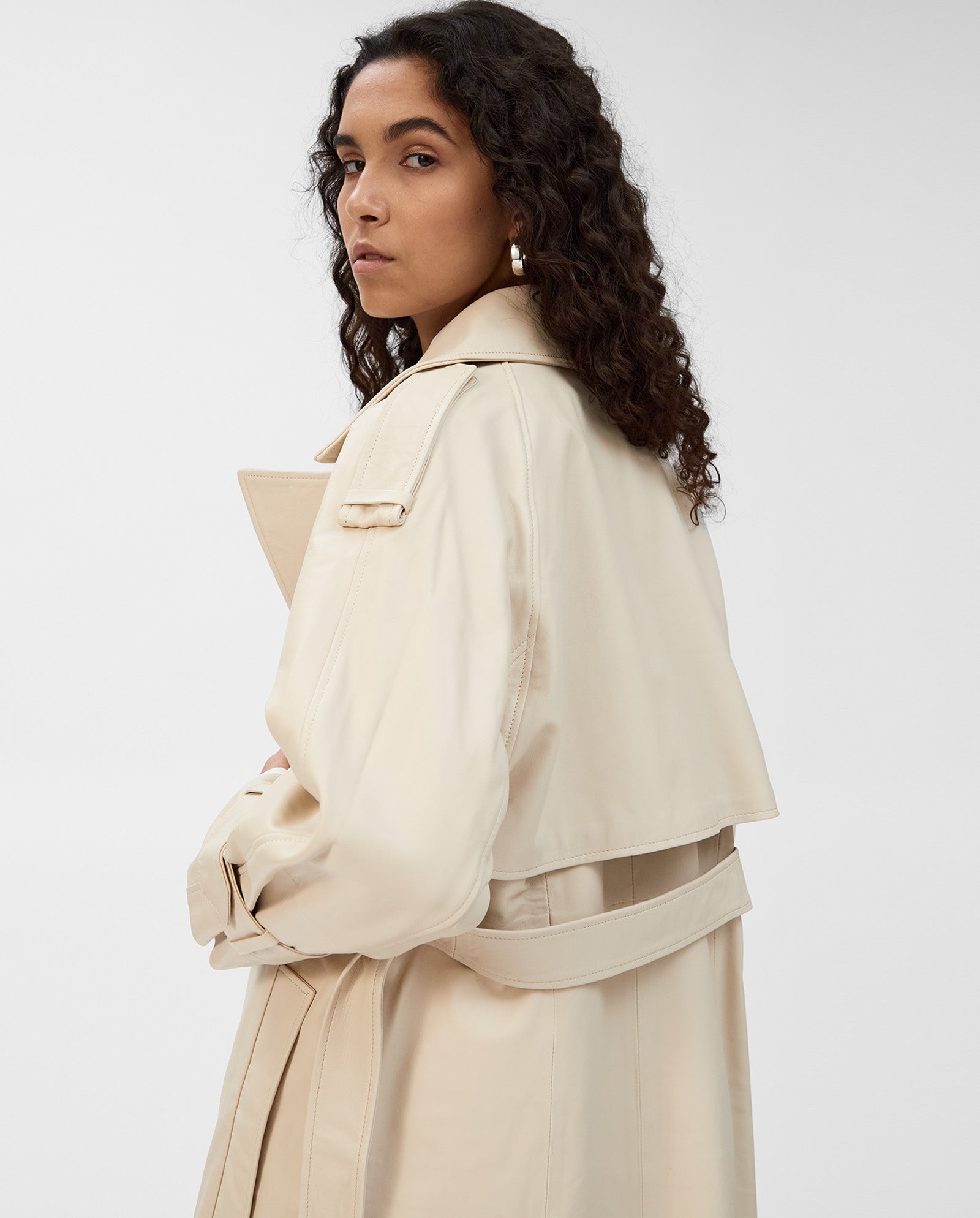 Woman with curly hair wears the IVY OAK LILITH Coat, a classic light beige trench with storm flap, looking over her shoulder against a plain background.