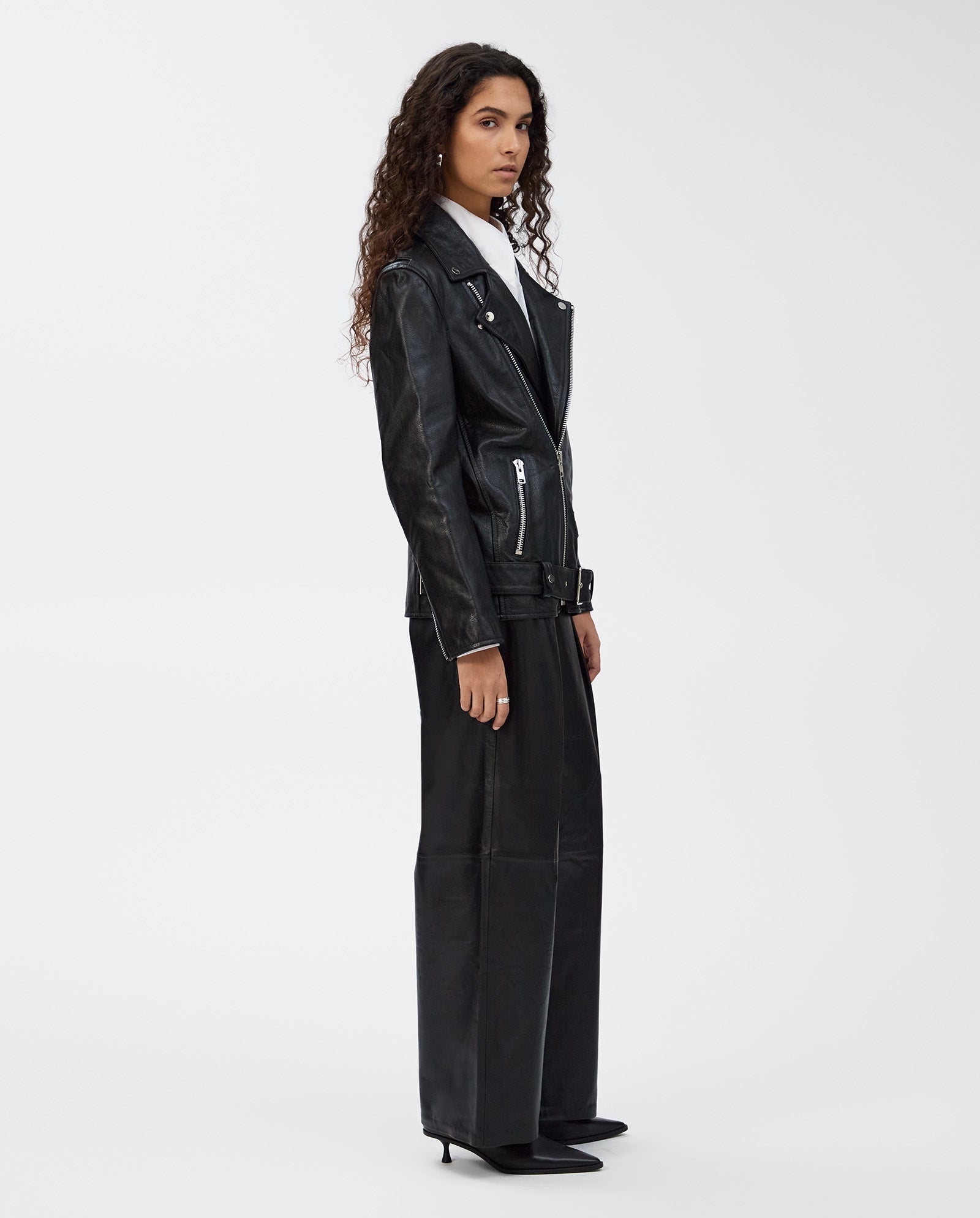 A woman wears the IVY OAK LENNY Jacket in black leather and wide-leg pants, standing sideways against a plain white background.
