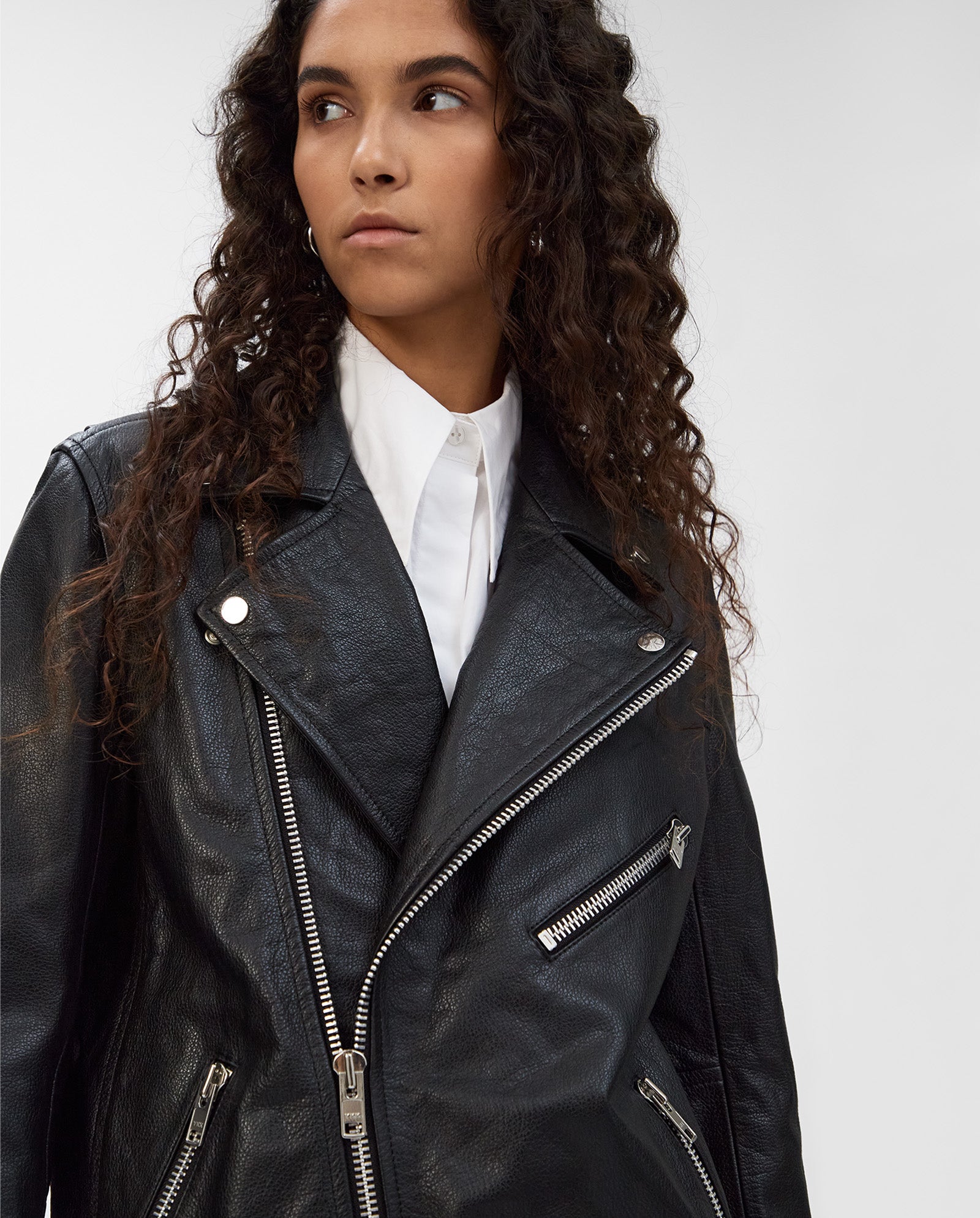 A woman with long curly hair wears the IVY OAK LENNY Jacket, an oversized black leather piece styled over a white collared shirt for a cool, relaxed look as she gazes to the side.