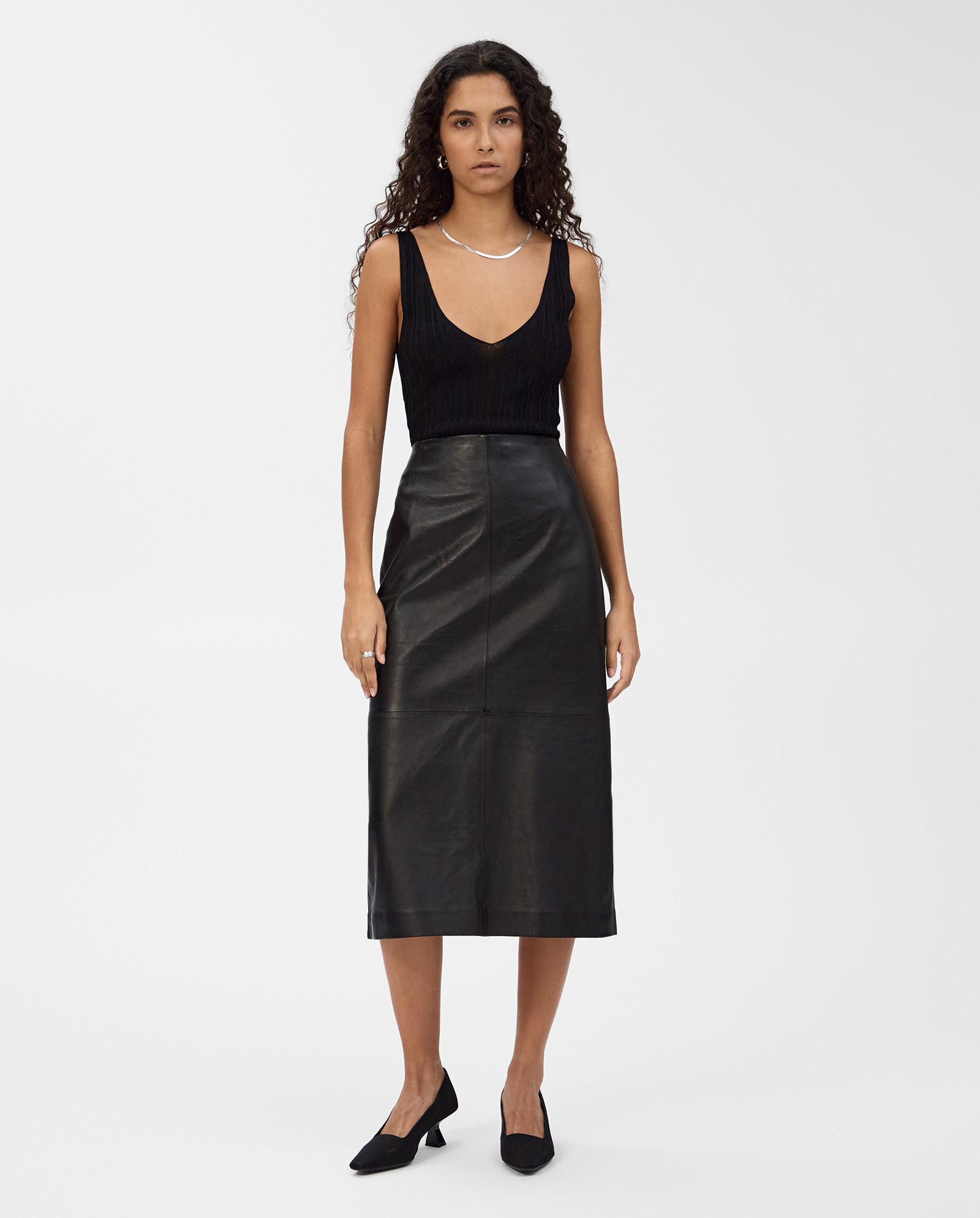 A woman wears the IVY OAK LEXIE Skirt, a high-waisted black leather midi skirt, paired with a black sleeveless top and black shoes, standing against a white background.
