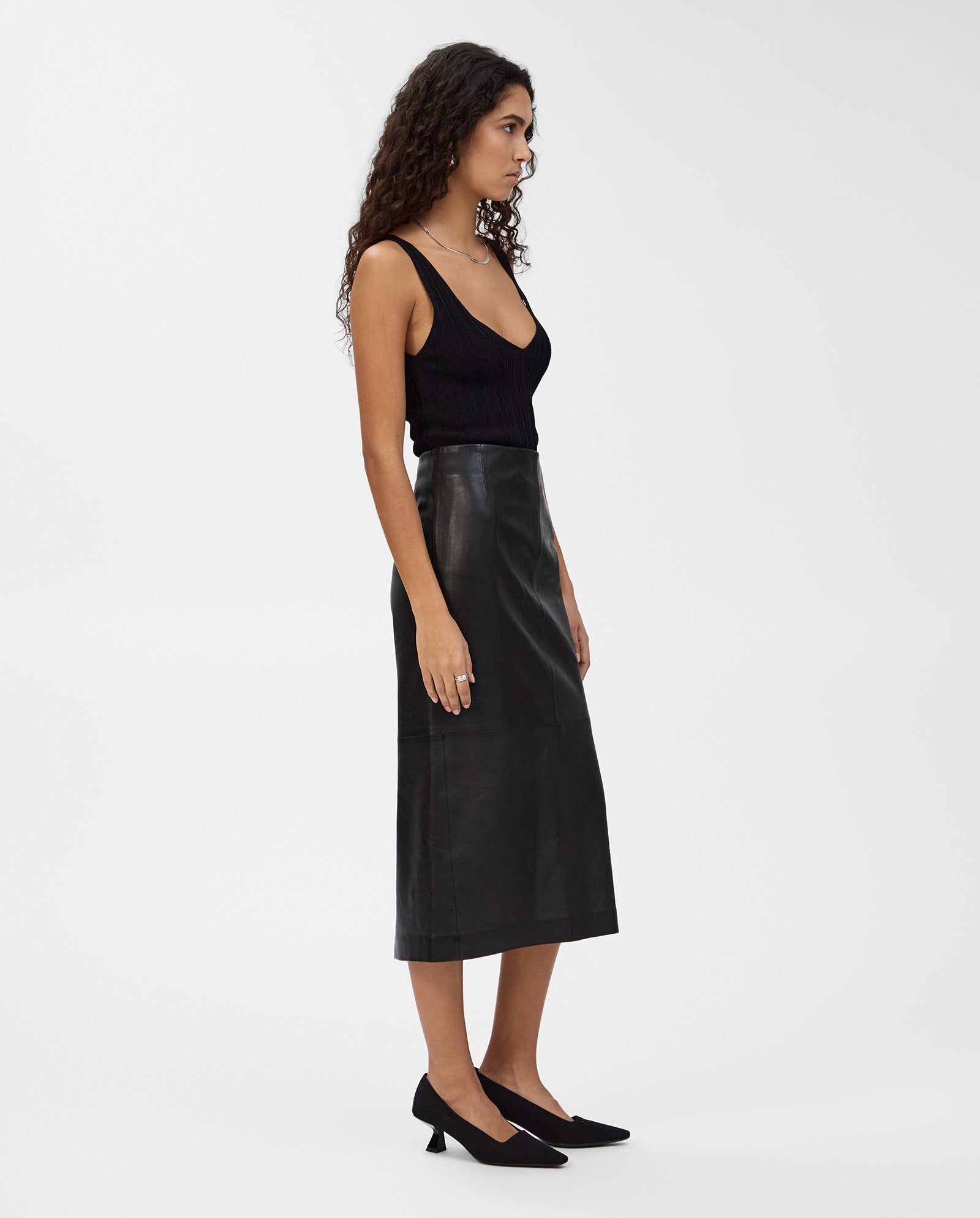 A woman stands sideways in black heels and a sleeveless top, wearing the IVY OAK LEXIE Skirt—a high-waisted, black, midi-length leather piece—against a white background.