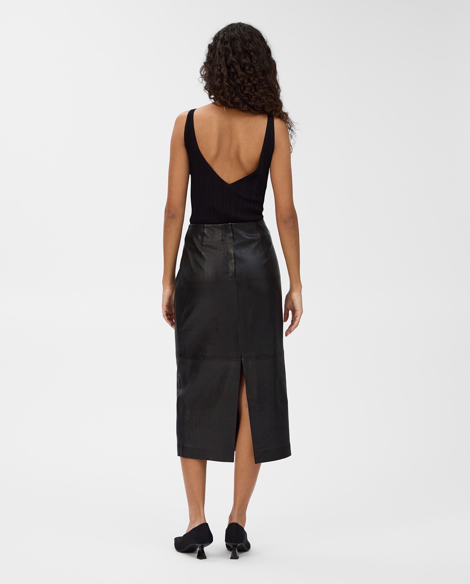 Woman wearing the IVY OAK LEXIE high-waisted black leather midi skirt with a back slit, paired with a black sleeveless top, standing facing away.