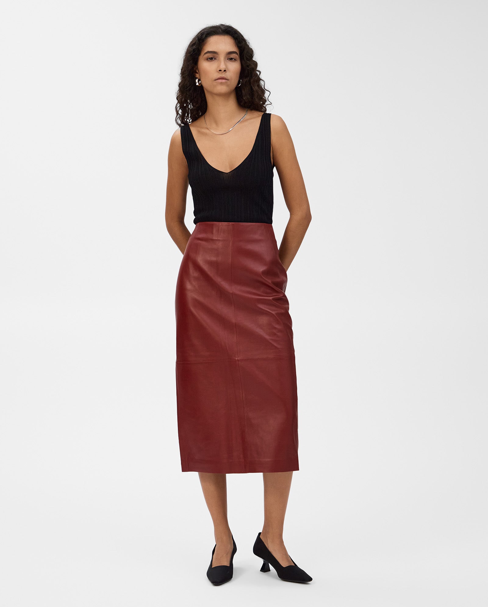 A woman wears the IVY OAK LEXIE Skirt, a high-waisted burgundy leather midi skirt, with a black sleeveless top and black heels against a white background.