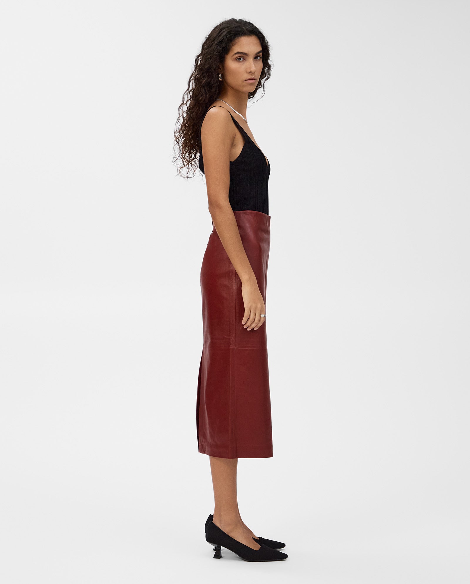 A woman stands in profile against a plain white background, wearing a black tank top and the maroon LEXIE Skirt by IVY OAK—a high-waisted leather midi skirt.