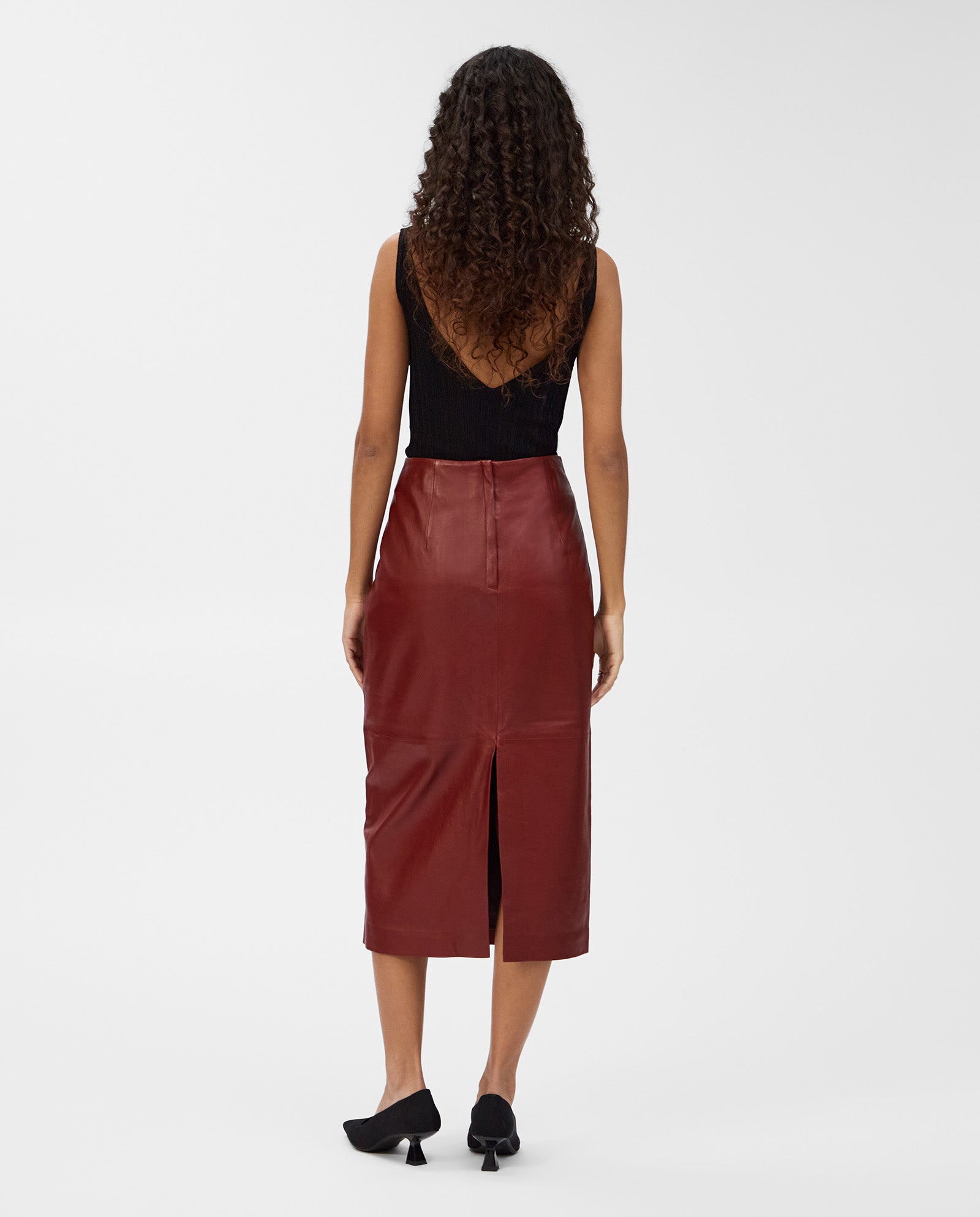 A woman with curly hair wears the IVY OAK LEXIE Skirt, a red high-waisted leather piece, paired with a black sleeveless top, standing with her back to the camera.