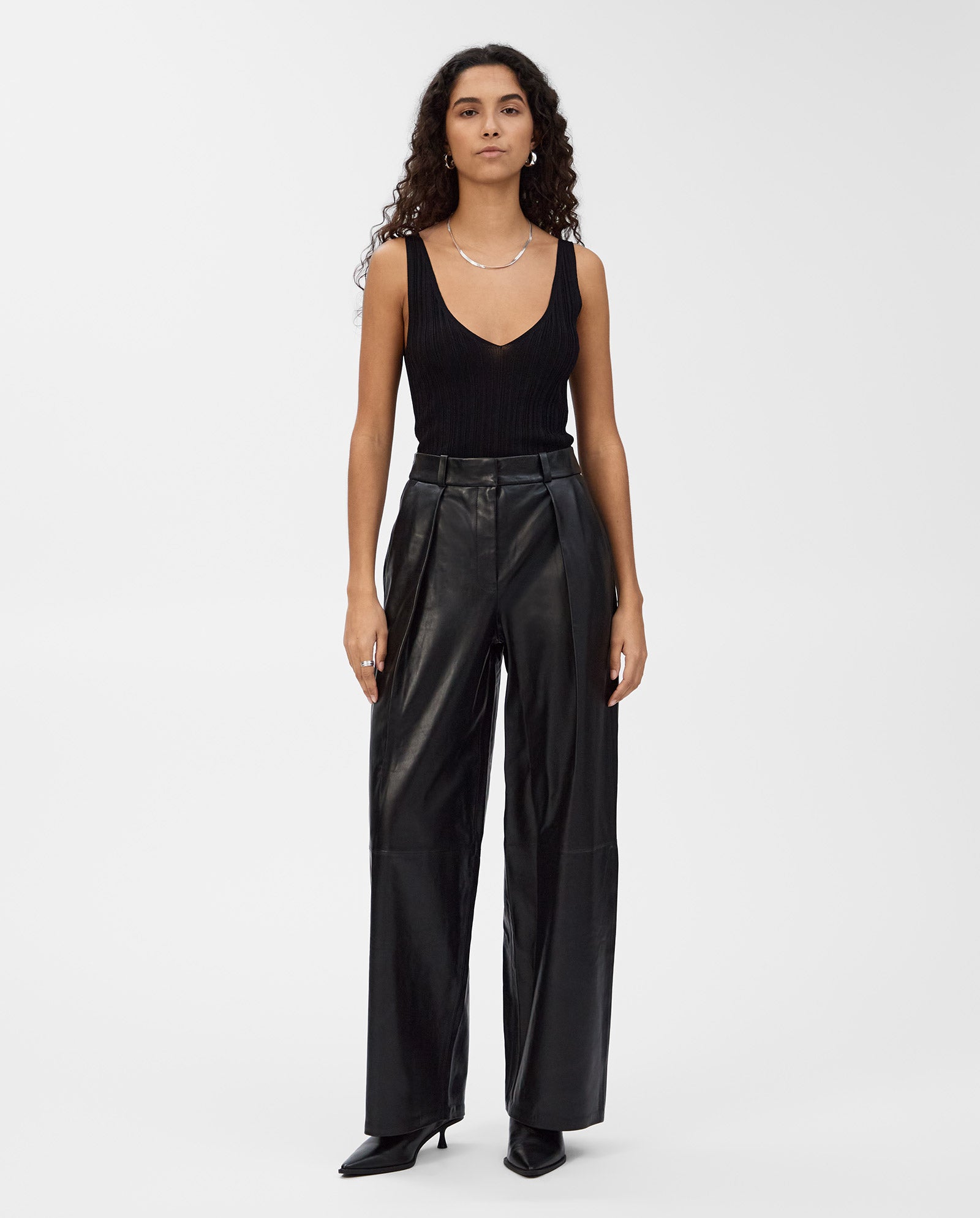 Woman wearing a black sleeveless top, IVY OAKs LORAINE classic cut leather trousers, and black pointed shoes, standing against a white background for a modern look.