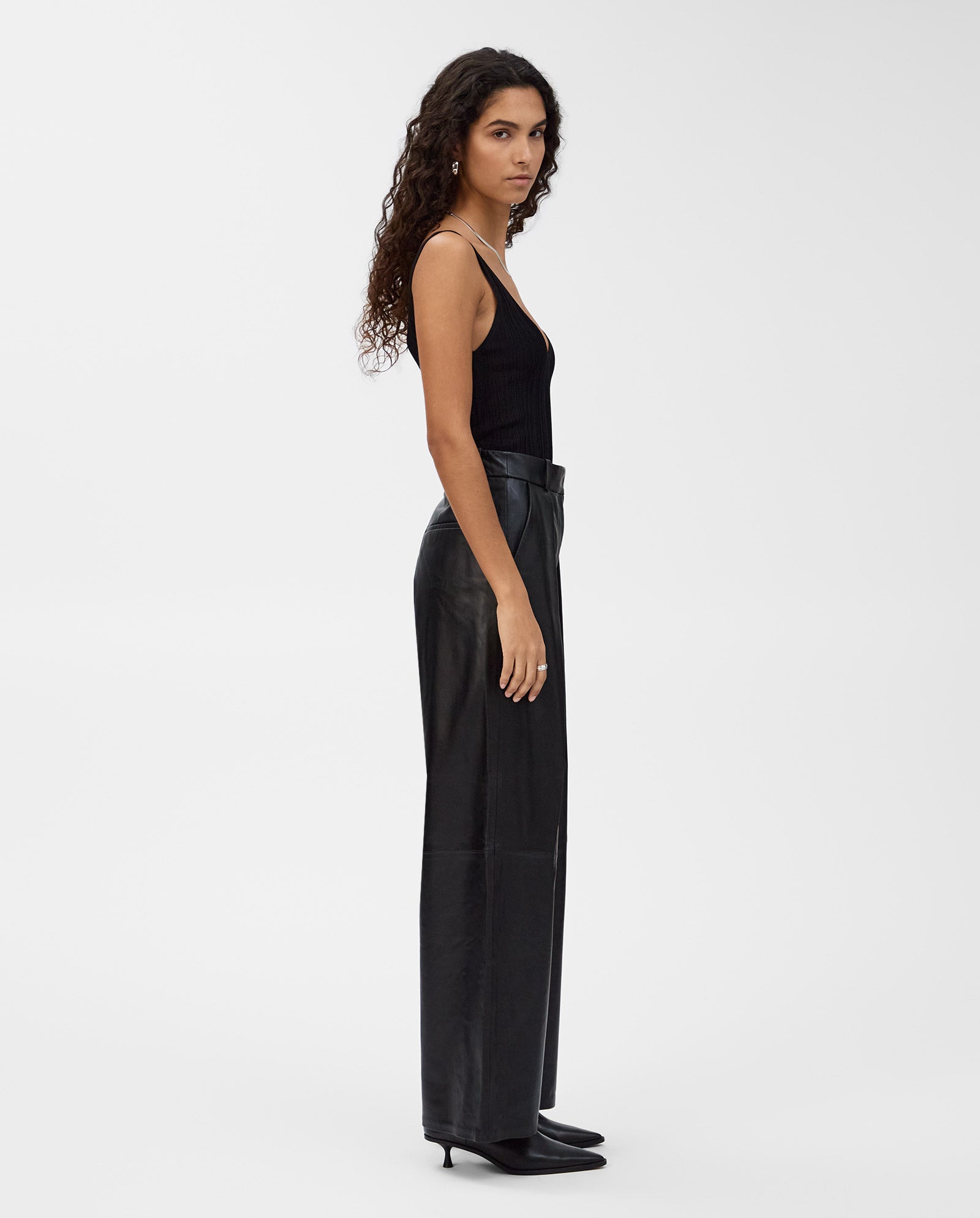 A woman with long curly hair wears a black tank top, IVY OAKs LORAINE Trouser in a classic cut, and black heels, standing sideways for a modern look.