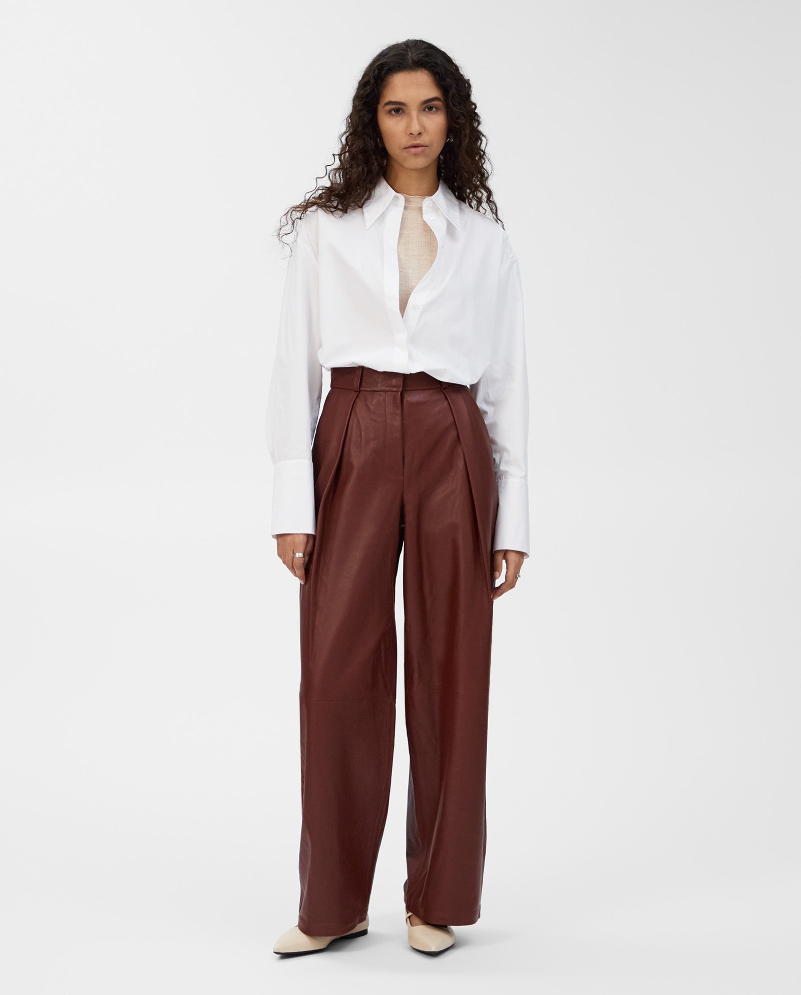 Woman in a white button-up shirt and IVY OAK LORAINE high-waisted, classic-cut brown leather trousers stands against a plain background for a chic modern look.