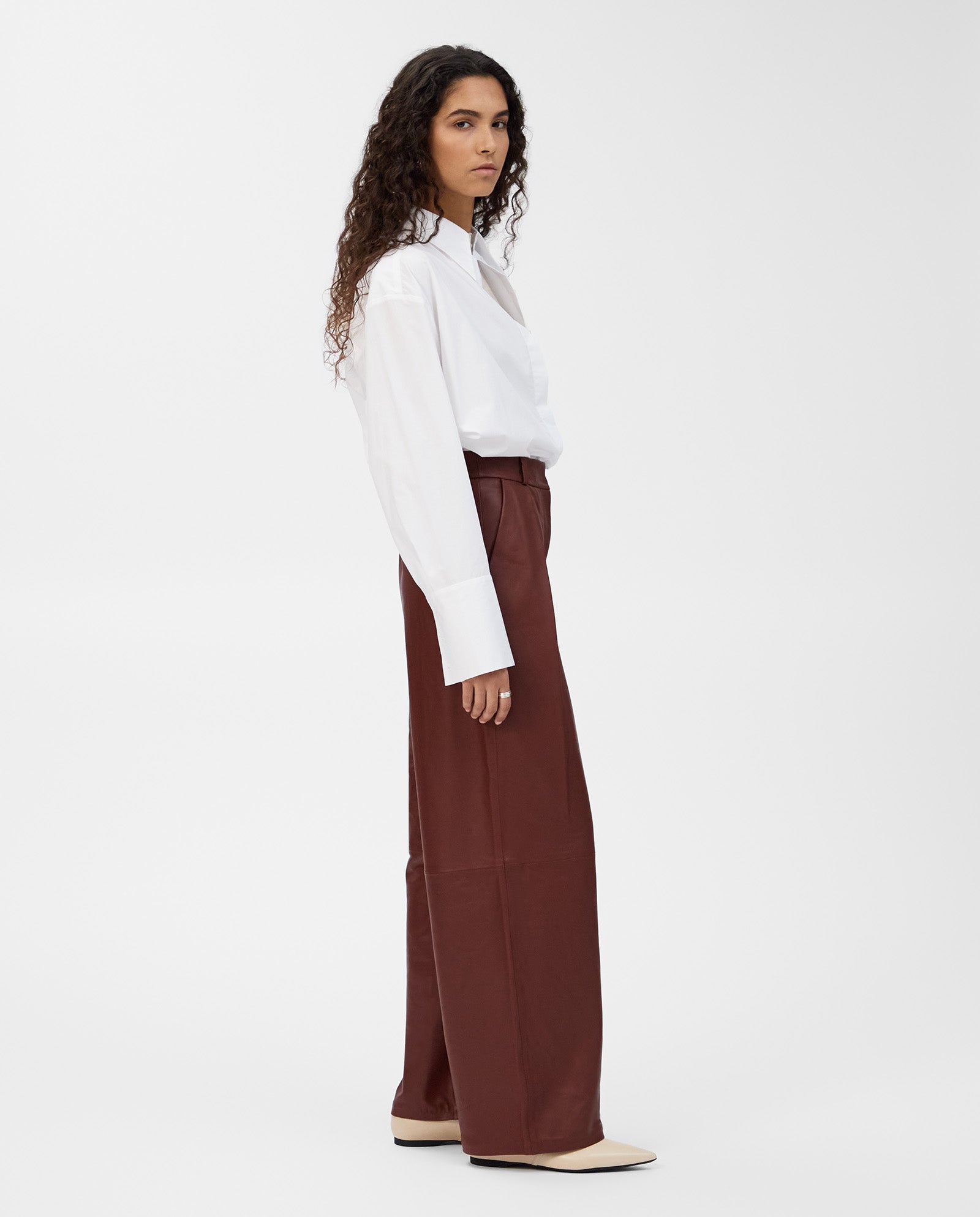 A woman stands sideways against a plain white background, wearing IVY OAK’s LORAINE Trouser in classic leather for a modern look.