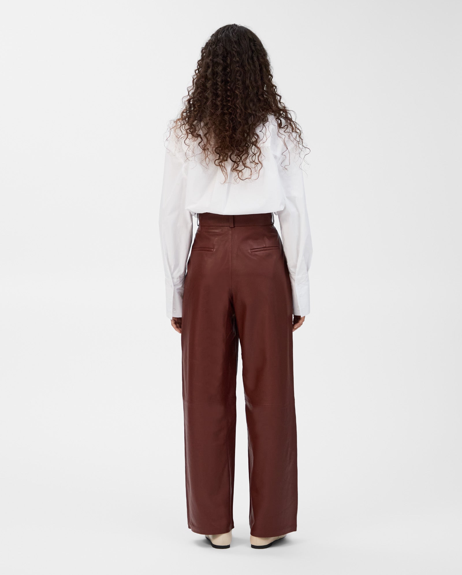 A person with long curly hair models the IVY OAK LORAINE Trouser in brown, paired with a white shirt, shown from the back for a modern look.