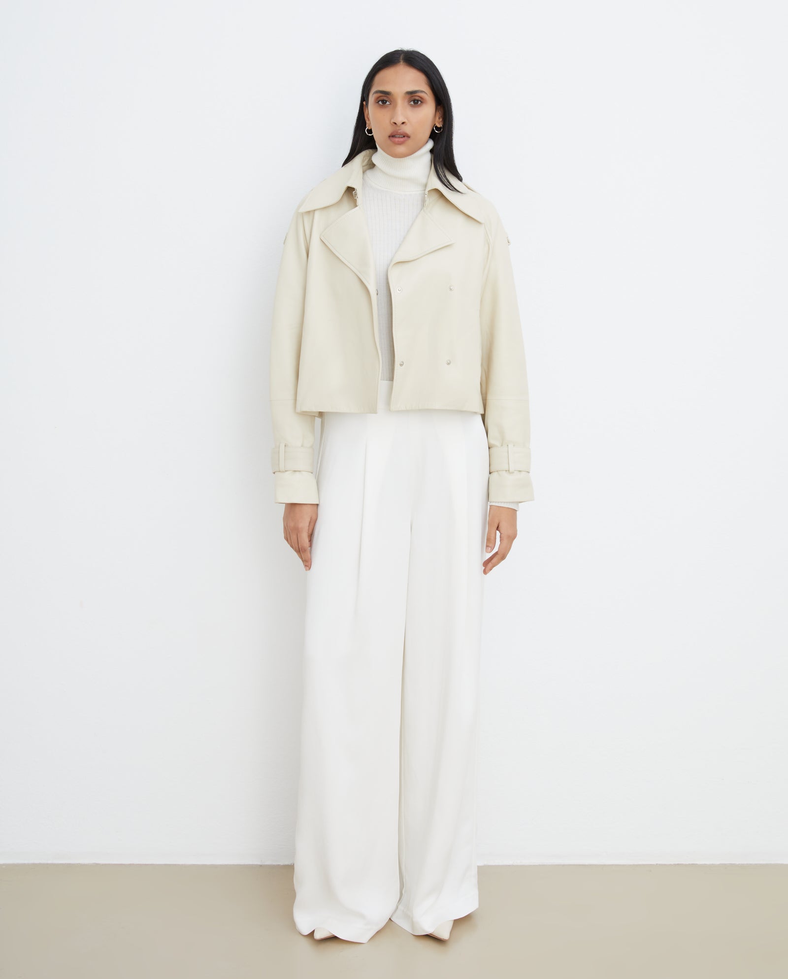 Woman stands against a white wall, wearing the IVY OAK LILITH ANN Jacket with a large collar, paired with a white turtleneck and wide-leg white pants.