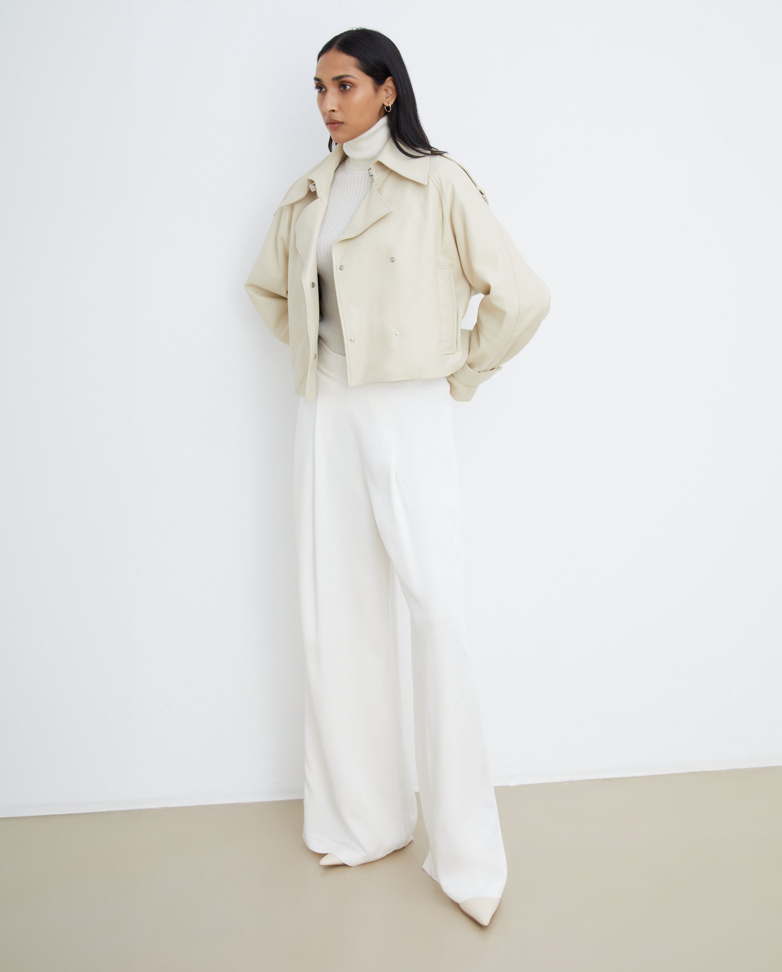 A woman wearing the IVY OAK LILITH ANN Jacket with a large collar, paired with a white turtleneck and wide-leg white pants, stands against a plain white wall.