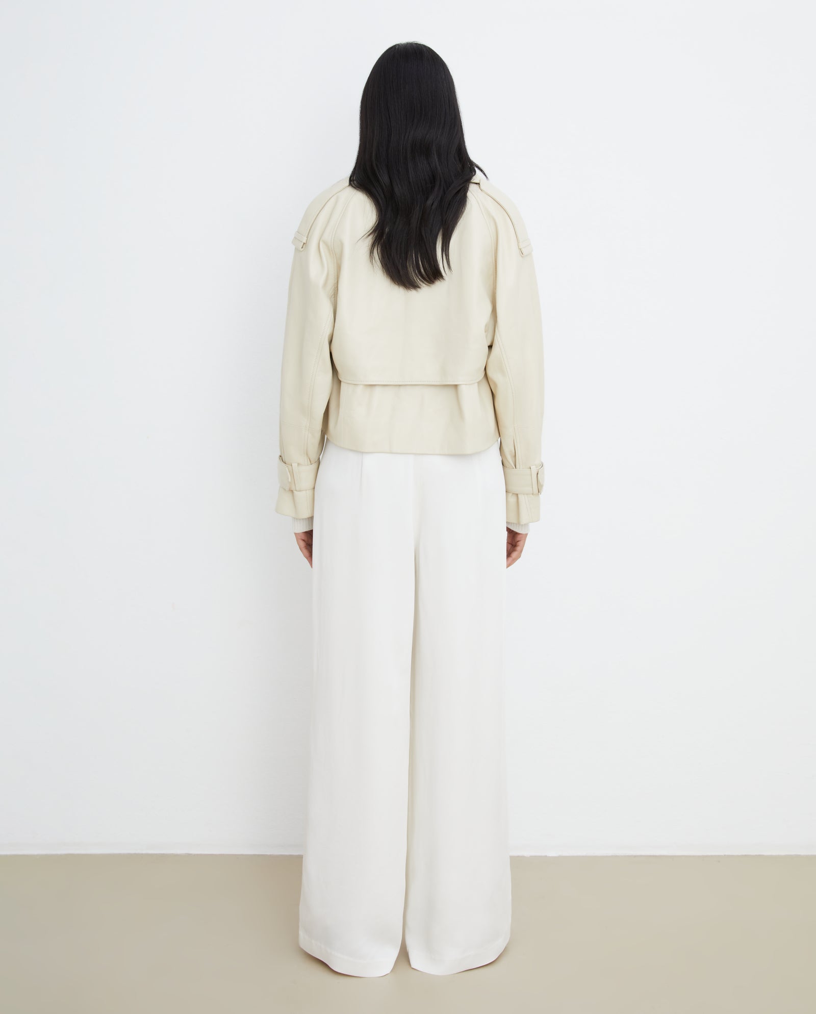 A person with long dark hair wears the IVY OAK LILITH ANN Jacket in cream, featuring a large collar, paired with wide-leg white pants, and stands facing a white wall.