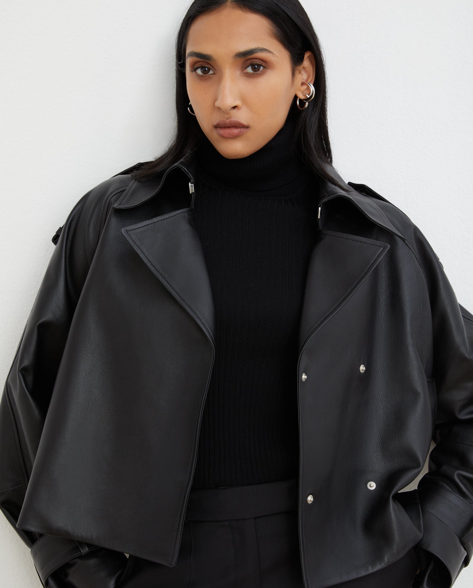 A woman with long dark hair wears the IVY OAK LILITH ANN Jacket—a black leather design with a large collar—styled over a black turtleneck, standing against a white wall.