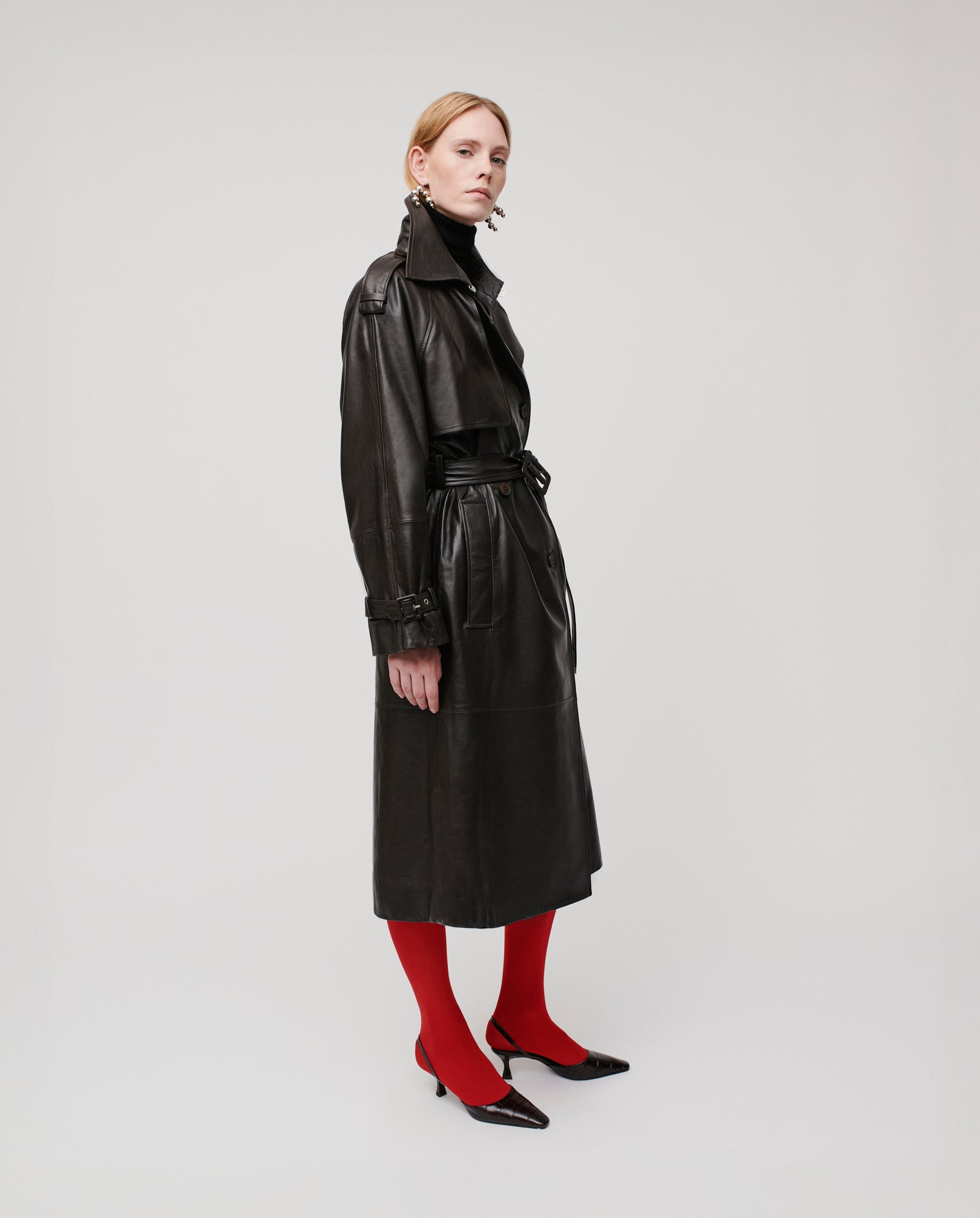 A woman wears the IVY OAK LINDA Coat, an oversized black leather trench, paired with red tights and black heels, standing against a plain background.