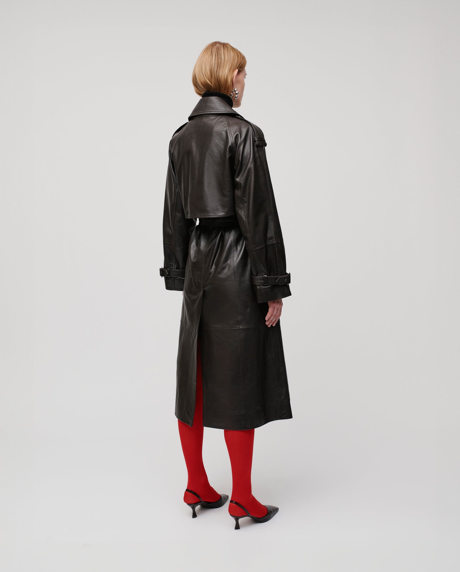 A person wears the IVY OAK LINDA double-breasted black leather trench coat, paired with red tights and black heels, standing facing away.