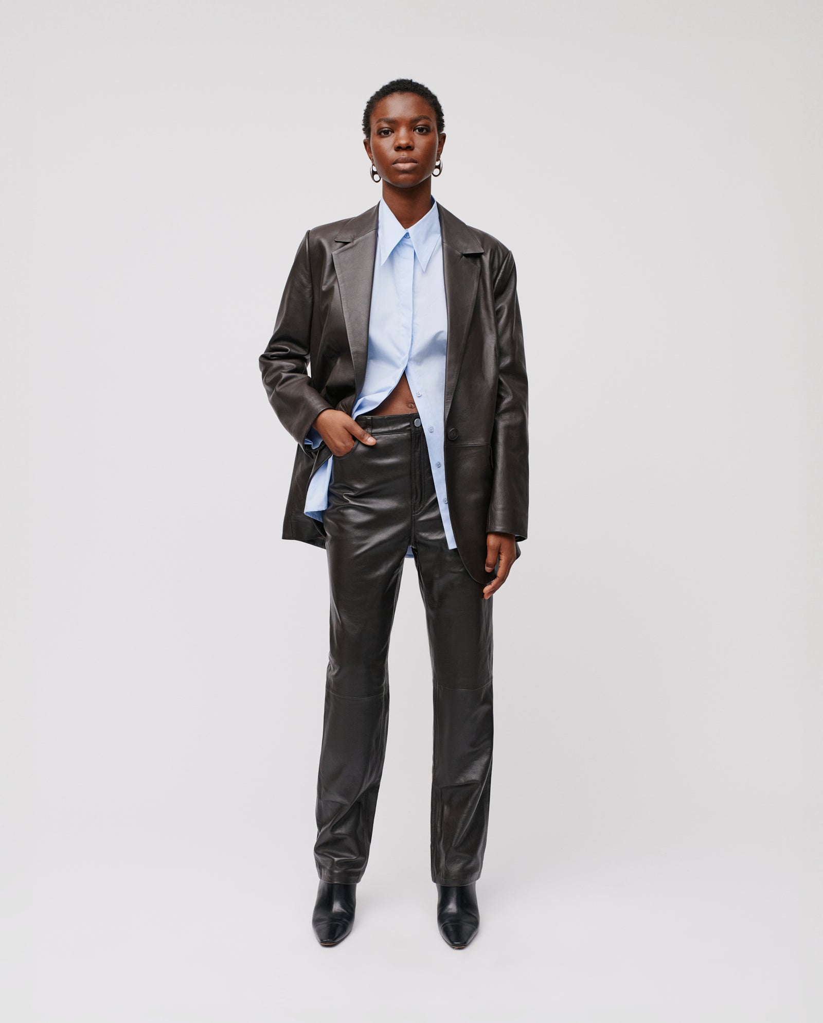 A person wears IVY OAKs LINDSEY Trousers, high-waisted with a straight leg, paired with a black leather jacket and light blue shirt, against a plain background.