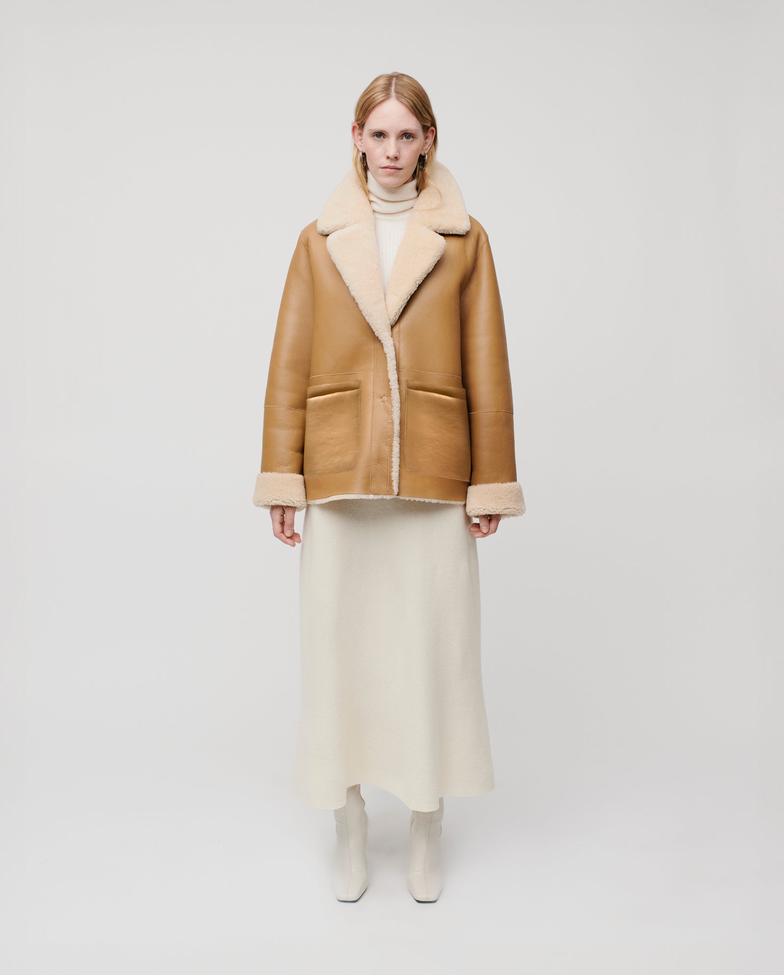 A woman in the IVY OAK LOTTE shearling jacket, cream turtleneck dress, and white boots stands against a plain background.