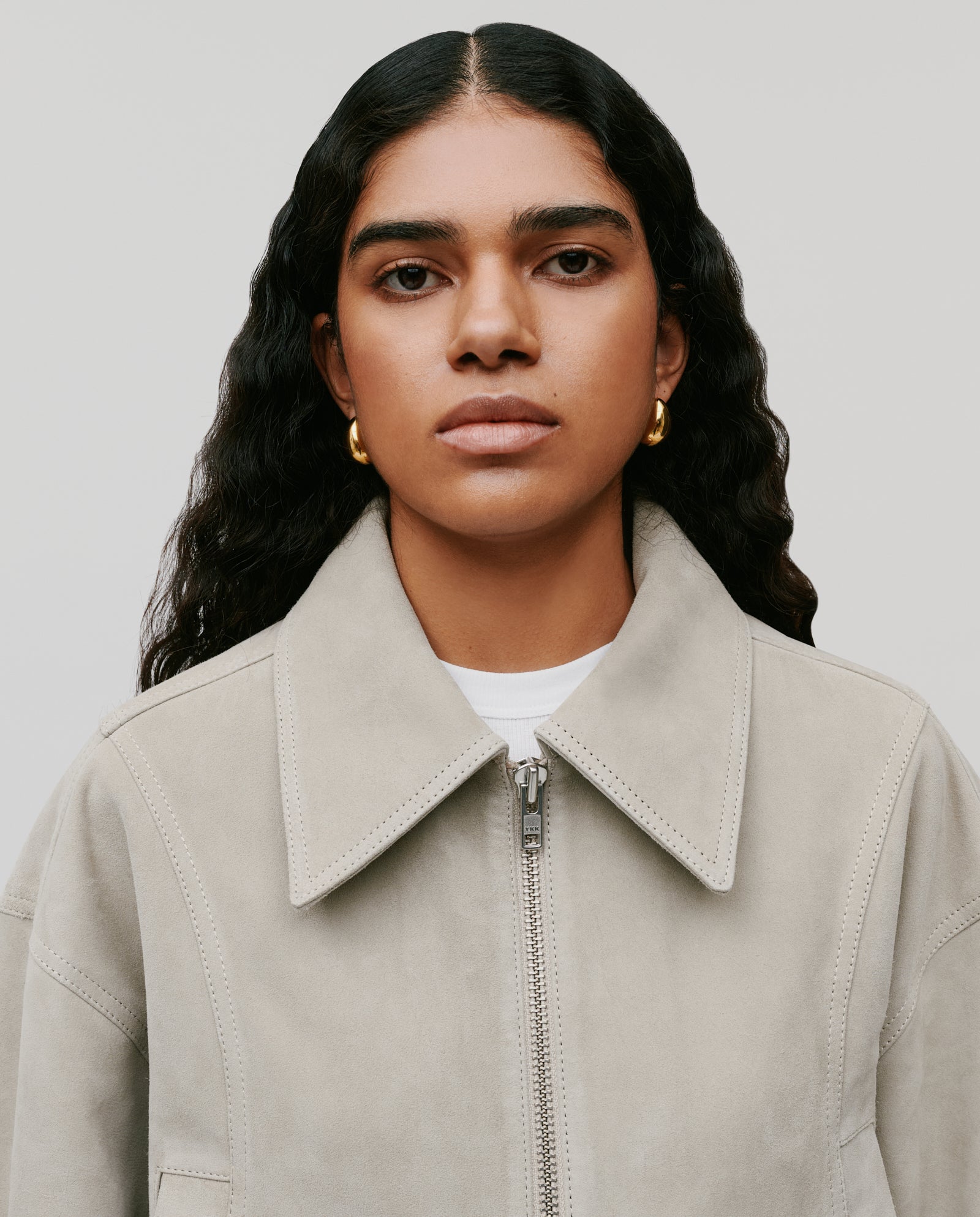 A woman with long wavy hair, facing forward, wears the IVY OAK LENA Jacket—a beige suede cropped bomber in a boxy style—paired with gold hoop earrings.