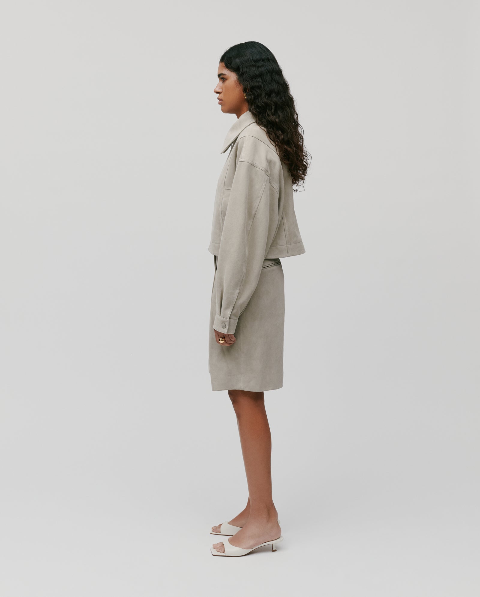 A woman stands in profile wearing the IVY OAK LENA Jacket, a beige suede bomber with wide sleeves, styled with a matching skirt and white heels against a plain background.
