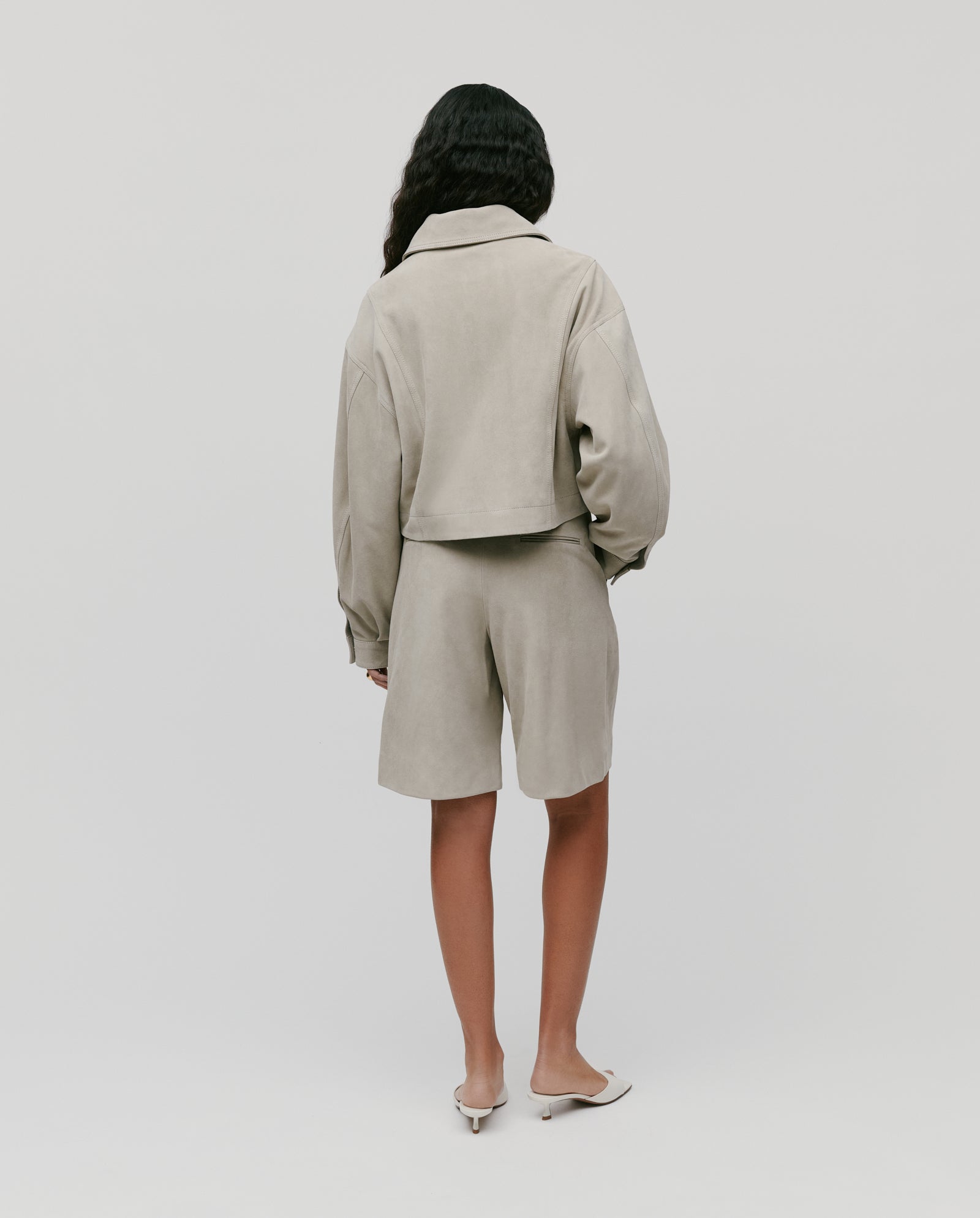 A woman wearing the IVY OAK LENA Jacket, a beige suede bomber with wide sleeves, and matching shorts stands facing away against a plain white background.