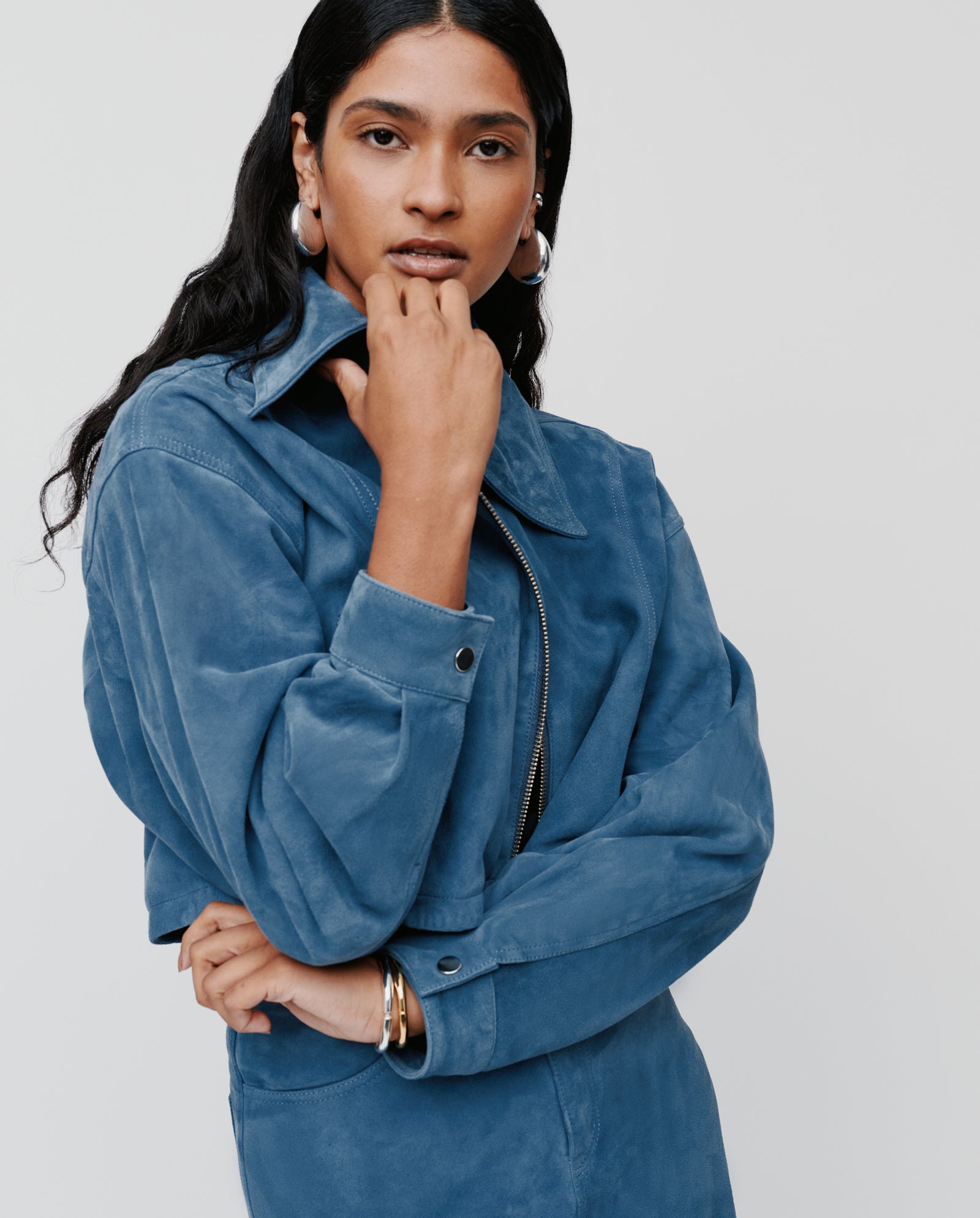A woman wearing the IVY OAK LENA Jacket, a blue suede bomber with wide sleeves, poses with her hand on her chin against a plain background.