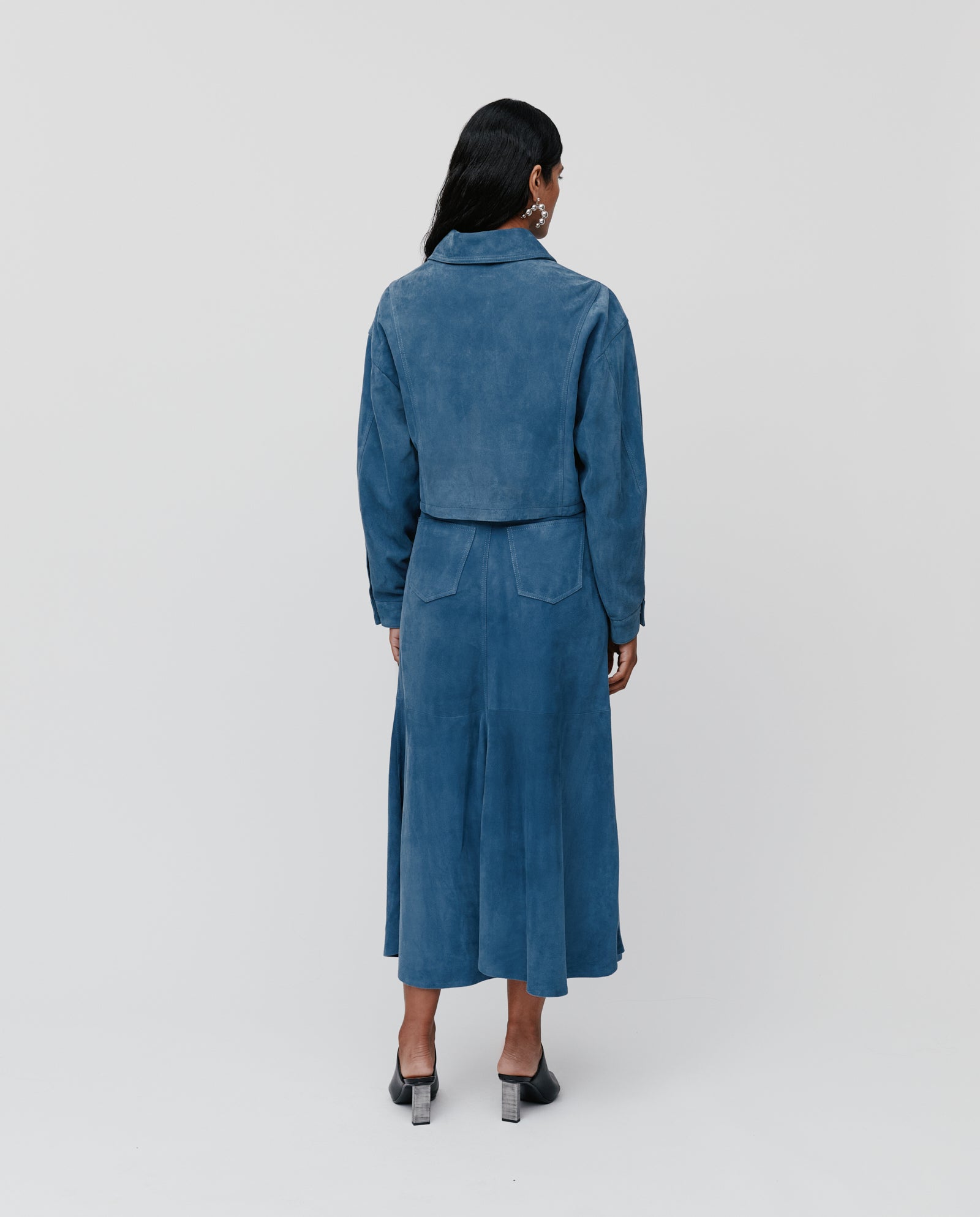 A person stands facing away against a plain background, wearing the IVY OAK LENA Jacket, a blue cropped boxy suede bomber with wide sleeves, paired with a matching skirt.