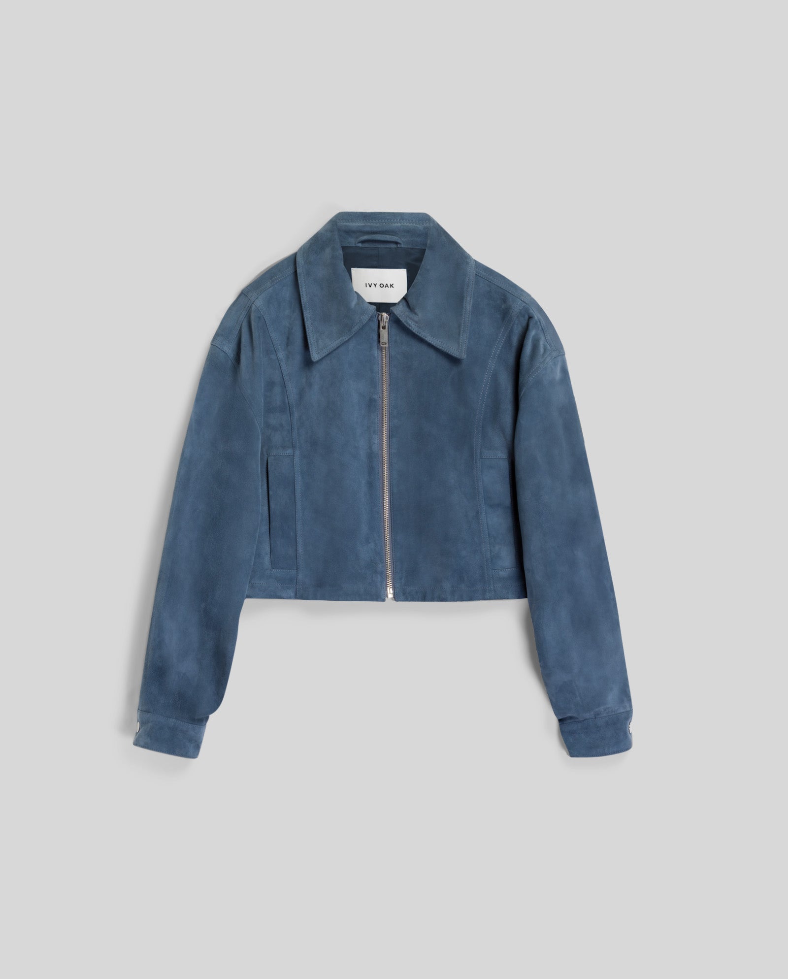 The LENA Jacket by IVY OAK is a blue suede bomber in a cropped, boxy fit with wide sleeves and a front zipper, shown on a plain light grey background.