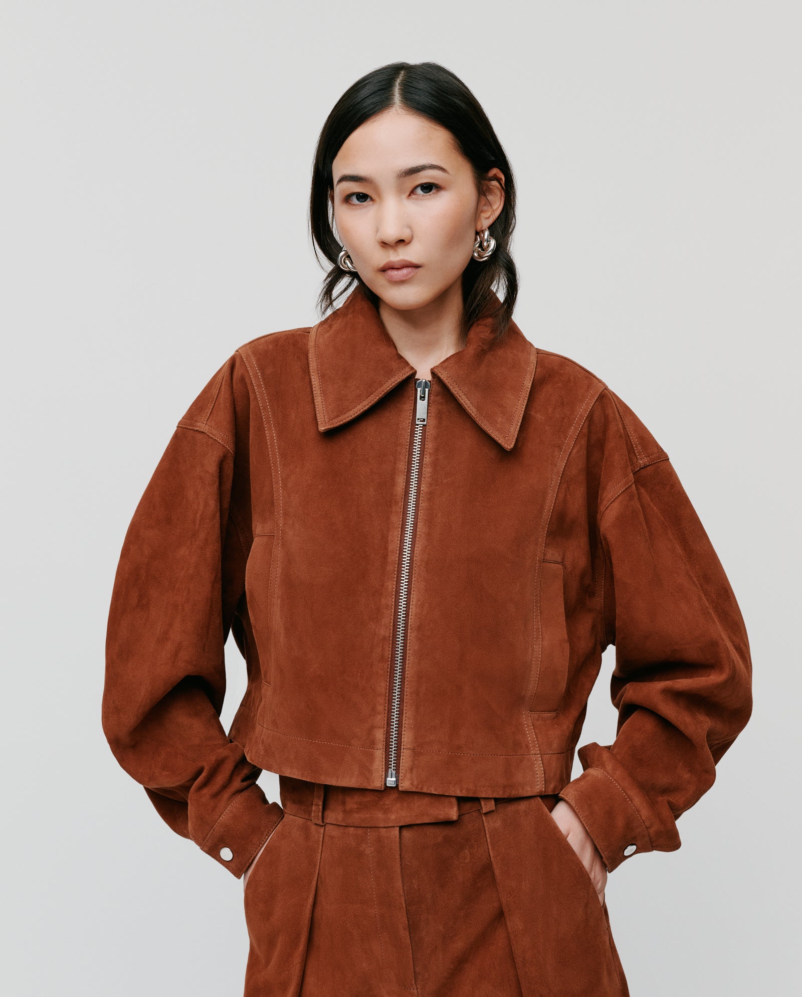 A woman wears the IVY OAK LENA Jacket, a brown suede bomber with wide sleeves, paired with matching pants. She stands with hands in pockets against a plain background.