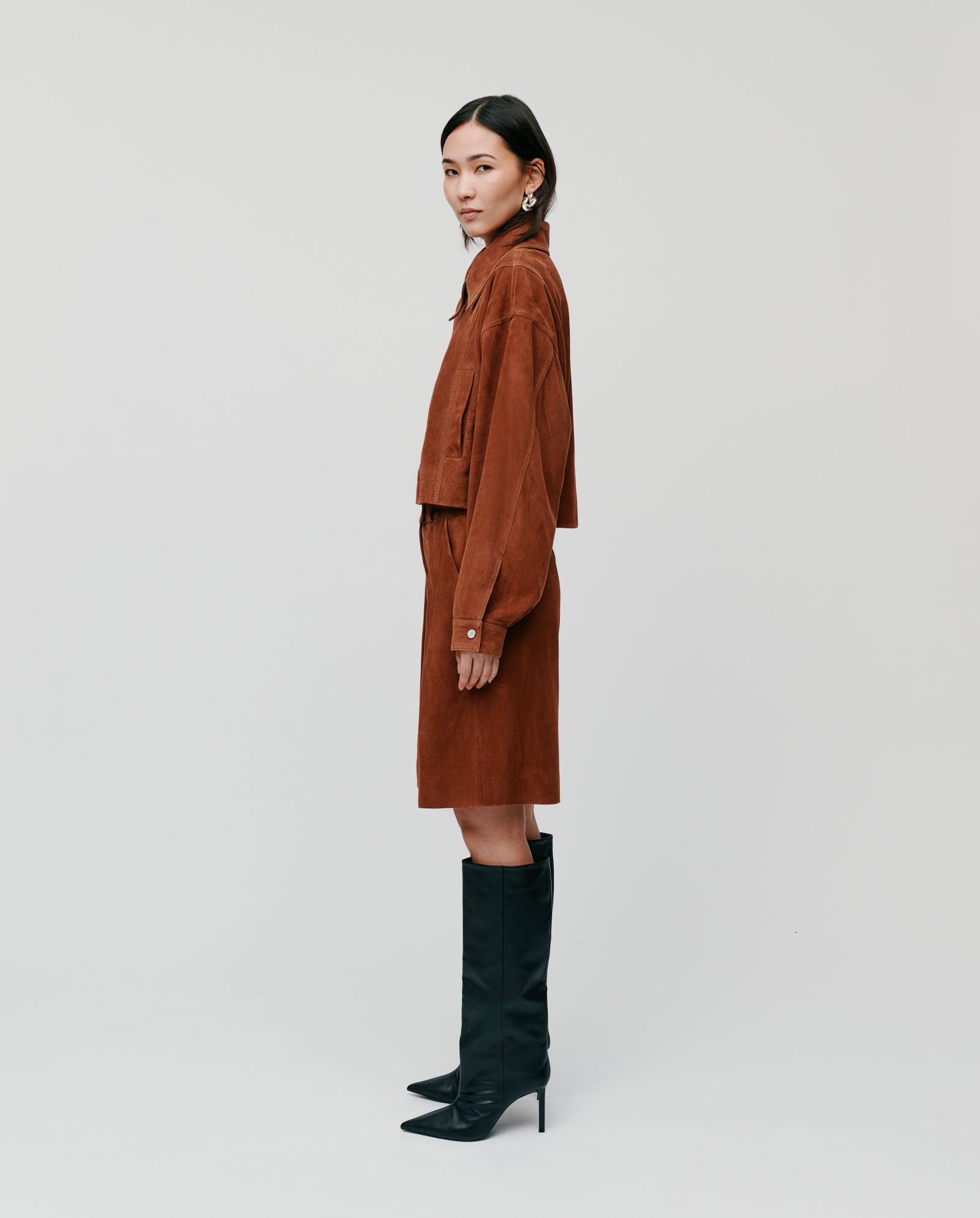 A woman wearing the IVY OAK LENA Jacket in brown suede, styled with a matching skirt and black knee-high boots, stands sideways.