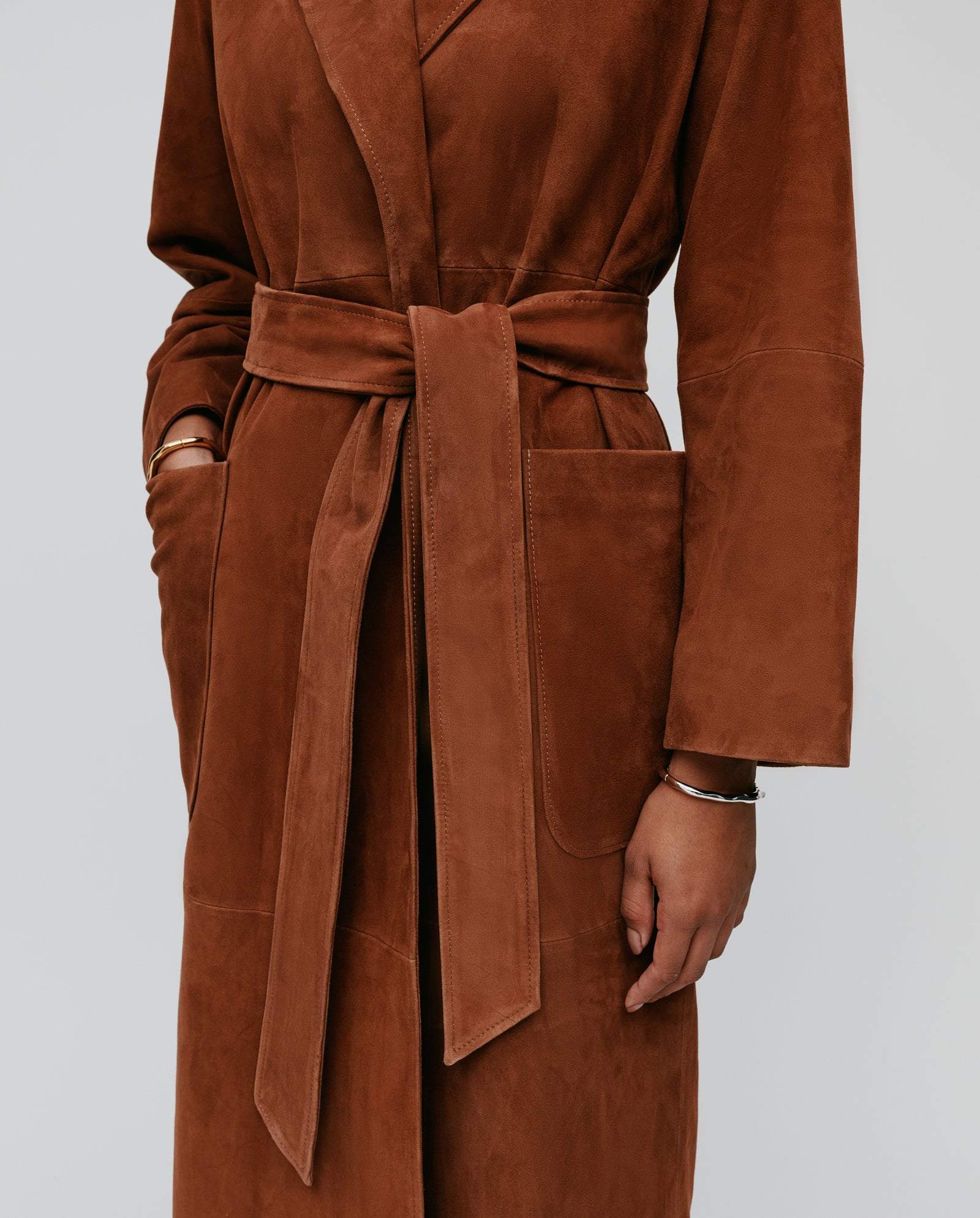 A person wears the LISA Coat by IVY OAK, a brown suede bathrobe-style coat with wide sleeves and hands partly in the pockets.