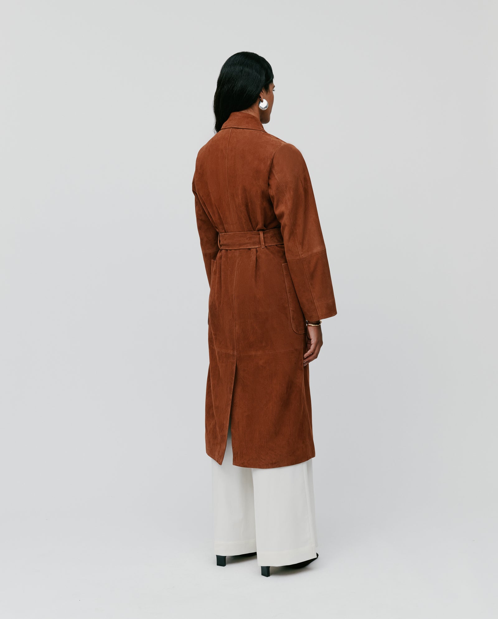 A person stands backwards against a plain background wearing the IVY OAK LISA Coat, a brown straight-cut suede coat with a belt, paired with white wide-leg pants.