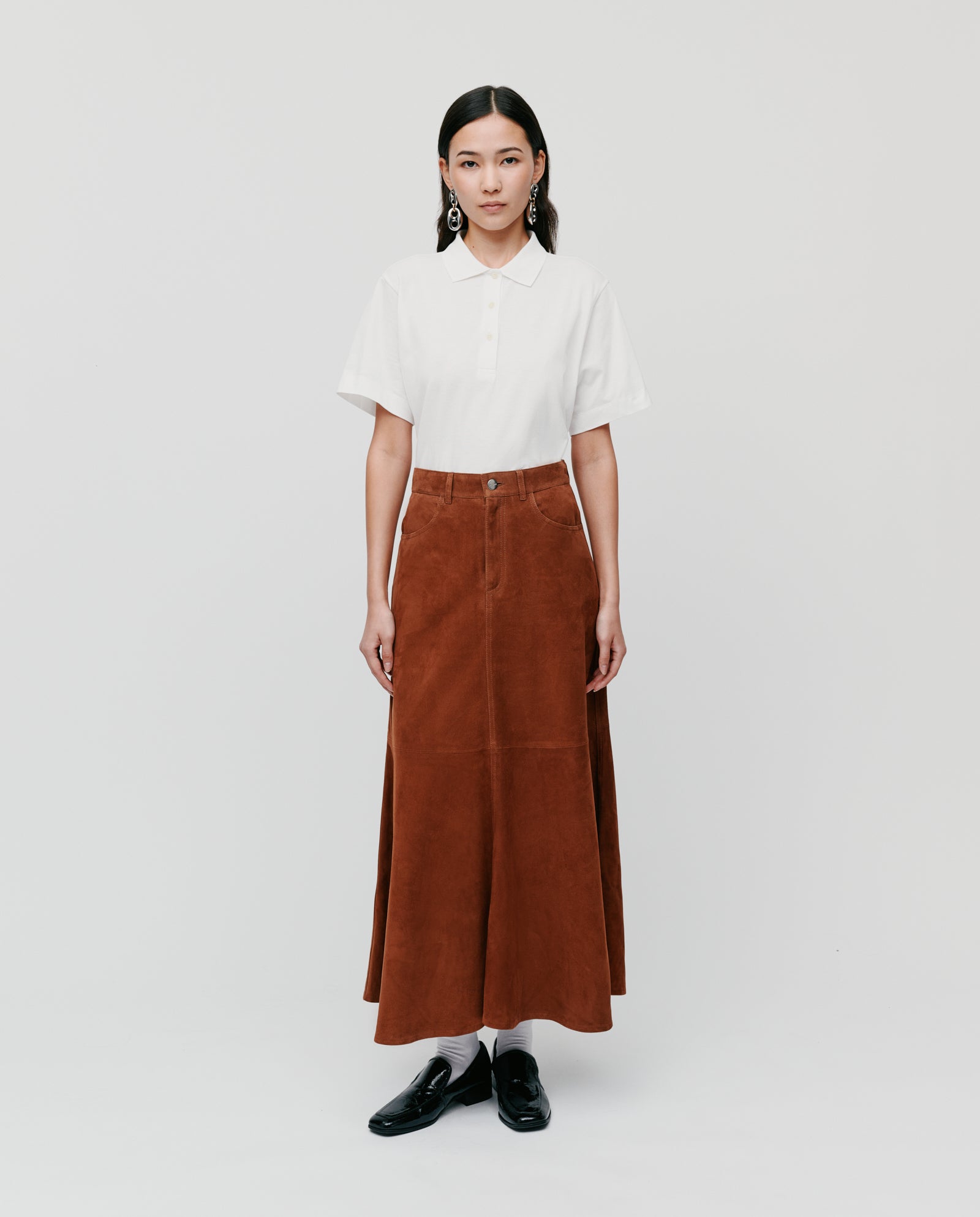 A woman stands against a plain white background wearing the IVY OAK LACY Skirt—a long, high-waist brown skirt—paired with a white polo shirt.