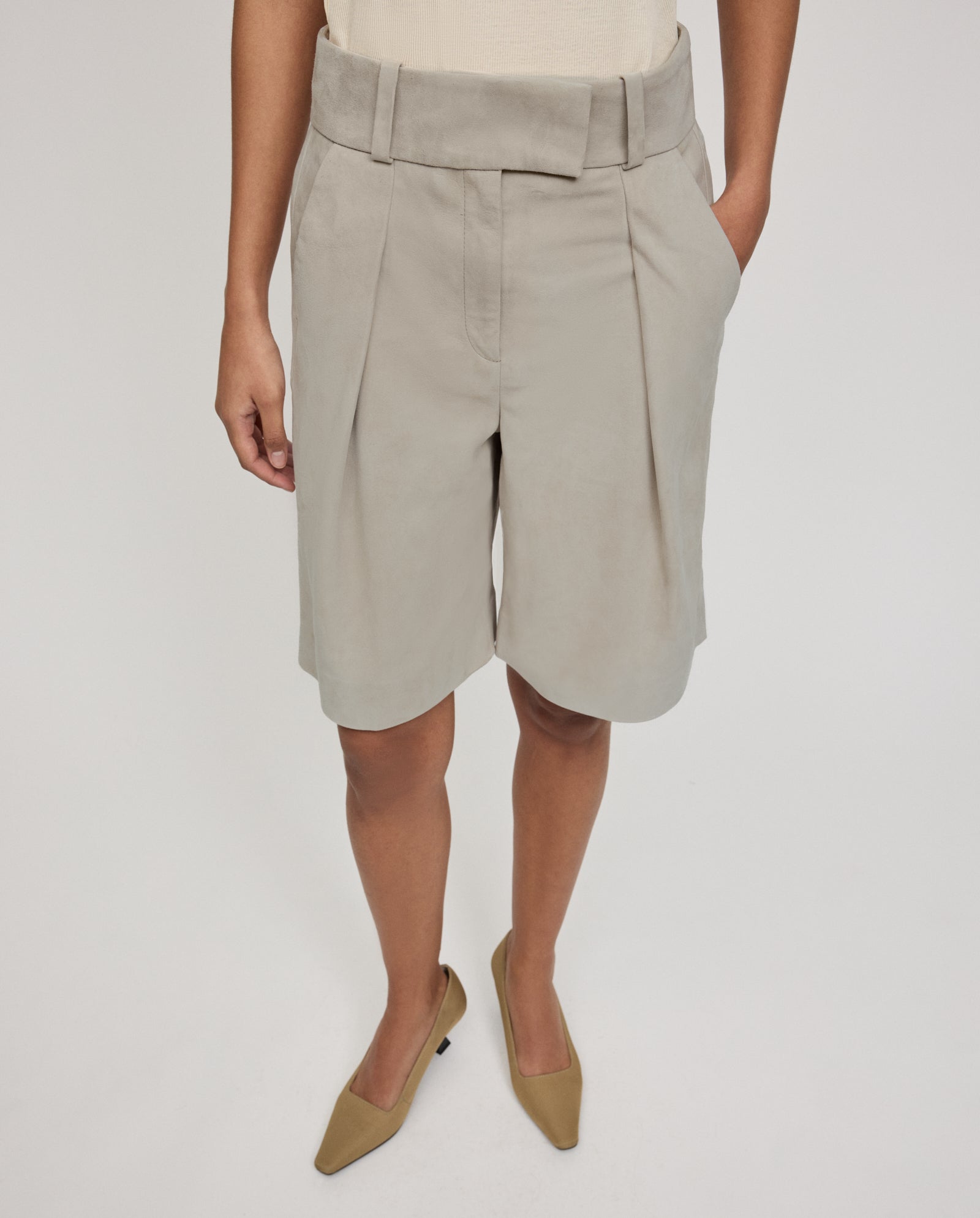 A person wearing IVY OAK’s LEA Trousers—high-waisted, light beige, pleated shorts—paired with taupe pointed-toe shoes, standing with one hand in their pocket.