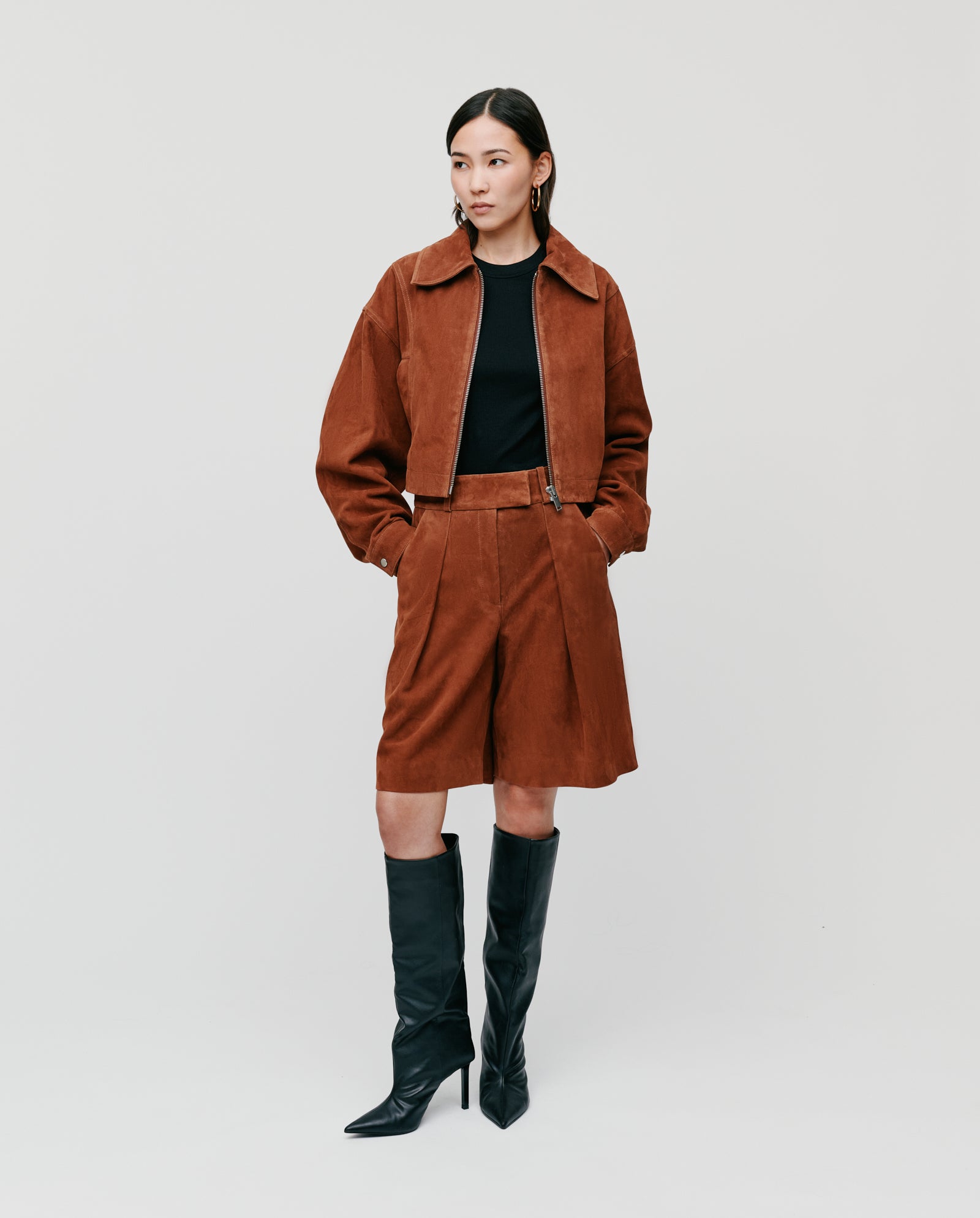 A woman wears IVY OAKs LEA Trousers—brown suede, high-waisted Bermuda shorts—paired with a black top and high black boots, standing against a plain light background.