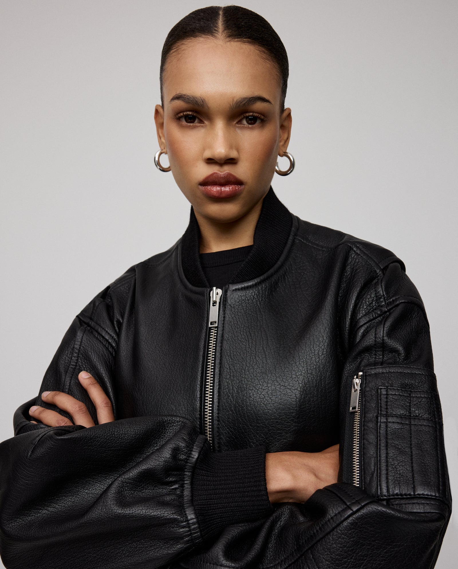 A woman with slicked-back hair and hoop earrings wears the IVY OAK LARA Jacket, a black premium hide leather design with a patched sleeve pocket. She stands with arms crossed and a neutral expression.