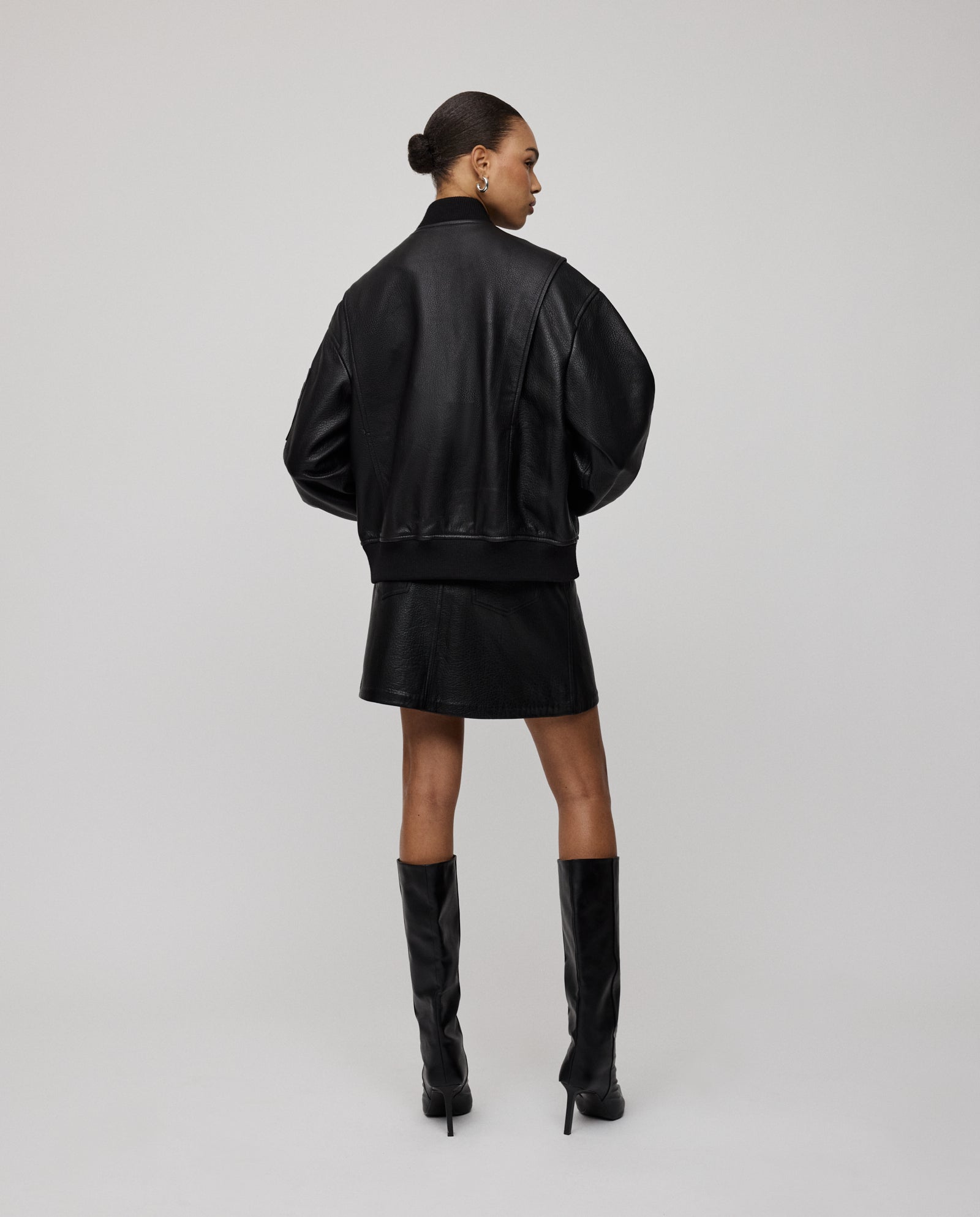 A woman faces away, wearing the oversized LARA Jacket by IVY OAK, paired with a matching skirt and knee-high black heeled boots.
