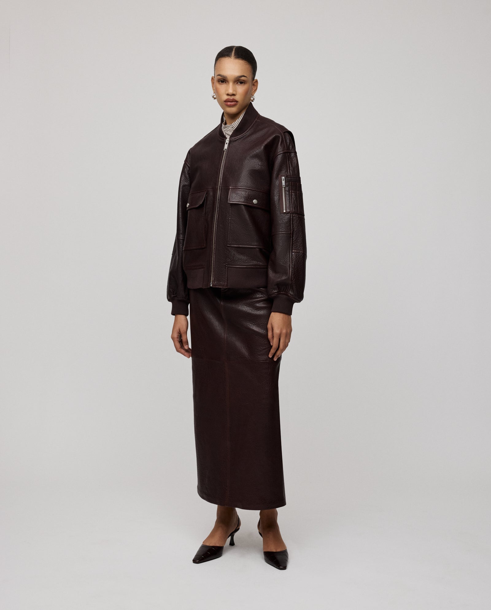 A woman models the IVY OAK LARA Jacket in premium dark brown leather with a patched sleeve pocket, paired with a matching long skirt and pointed heels, standing against a plain background.