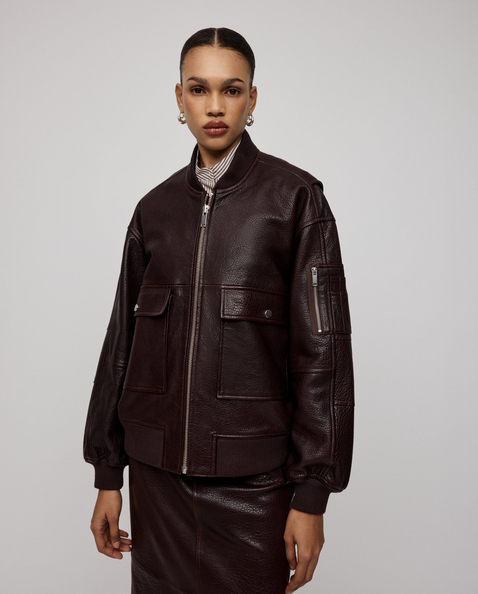 A woman wearing the IVY OAK LARA Jacket, a dark brown premium hide leather design with a patched sleeve pocket and skirt, stands against a plain light background.