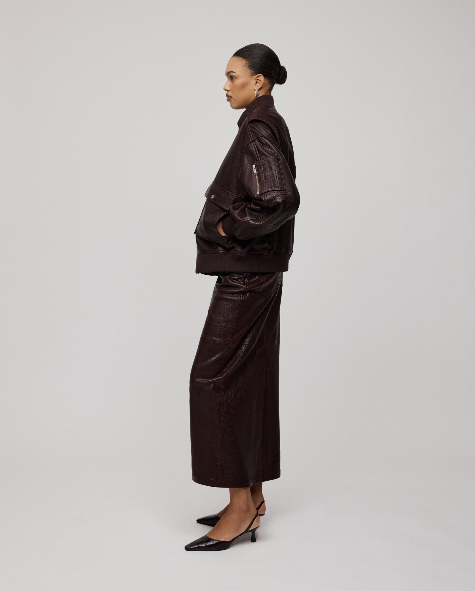 A woman in profile wears the IVY OAK LARA Jacket in premium dark brown leather with a patched sleeve pocket, styled with a skirt and black heels, standing against a plain background.