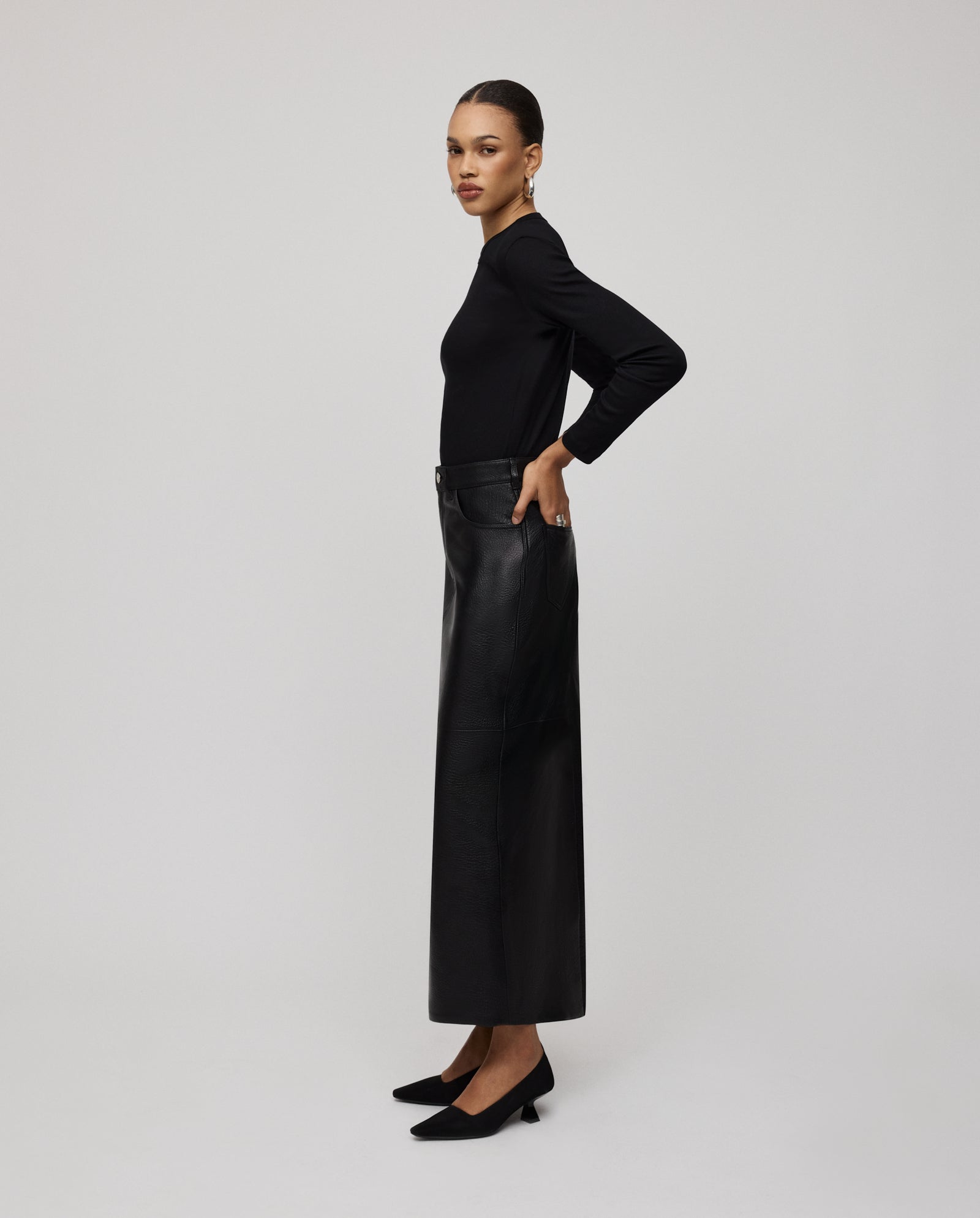 A woman stands sideways, wearing a black long-sleeve top, black shoes, and the IVY OAK LISANNE Skirt—a sleek, ankle-length piece with a flattering A-line silhouette.
