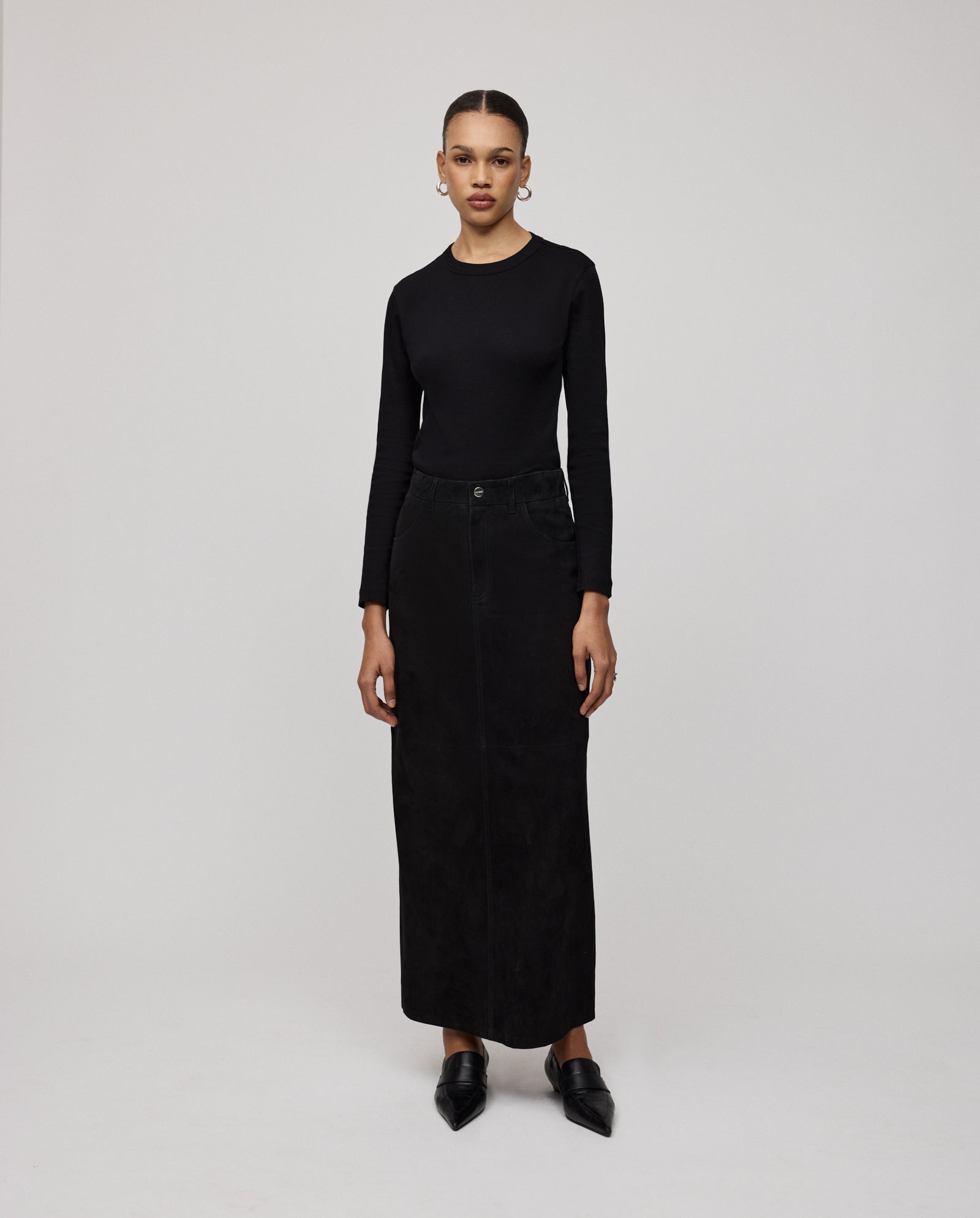 A woman stands against a plain background, wearing a black long-sleeve top, black shoes, and the IVY OAK LISANNA ROSE Skirt—a high waist A-line silhouette maxi skirt.