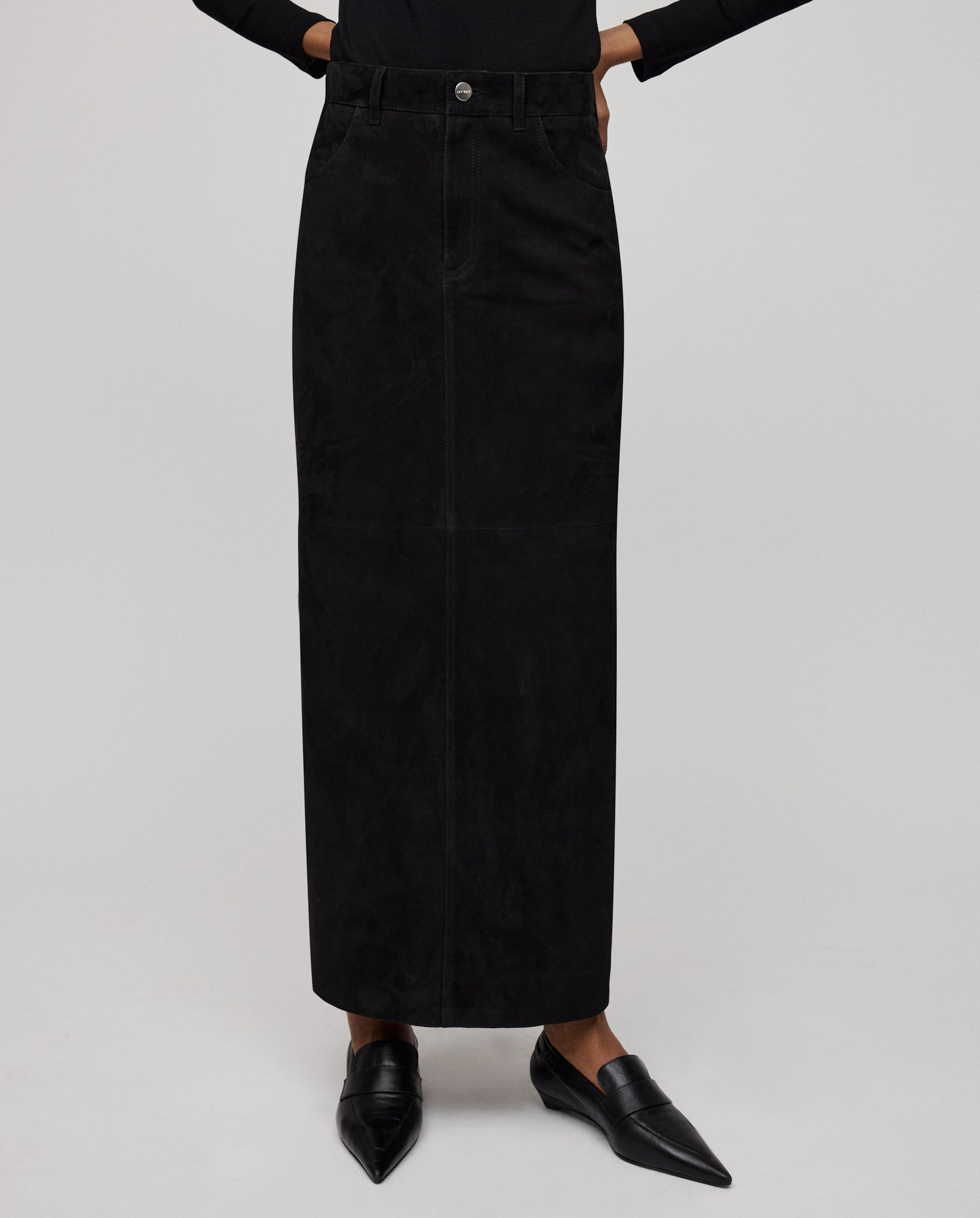 A person wears the IVY OAK LISANNA ROSE Skirt, a long black high-waist A-line design, paired with black loafers and a black top, standing against a plain background.