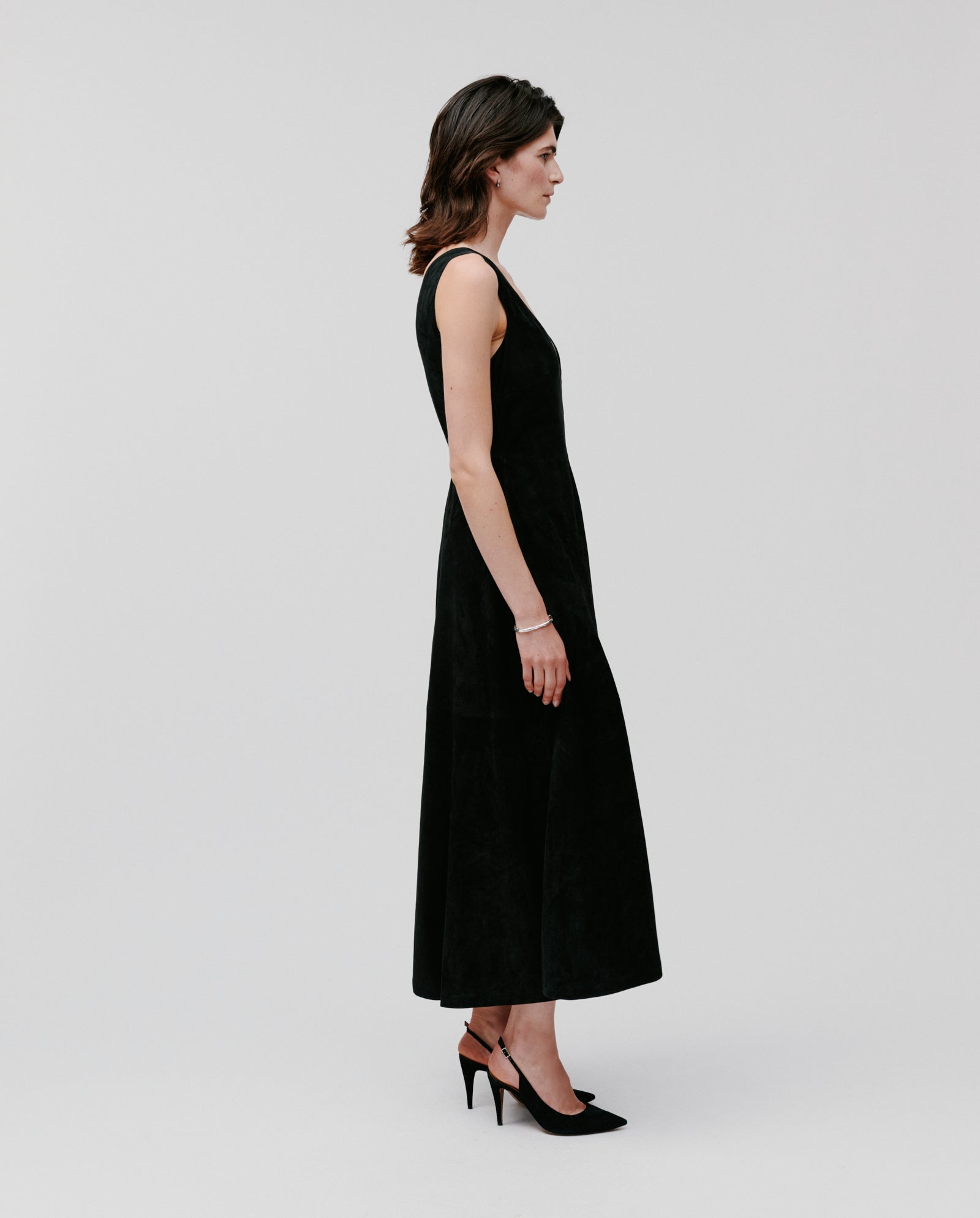A woman in a sleeveless, long black IVY OAK LORENA DRESS with a deep V-neckline and black heels stands in profile against a plain background.