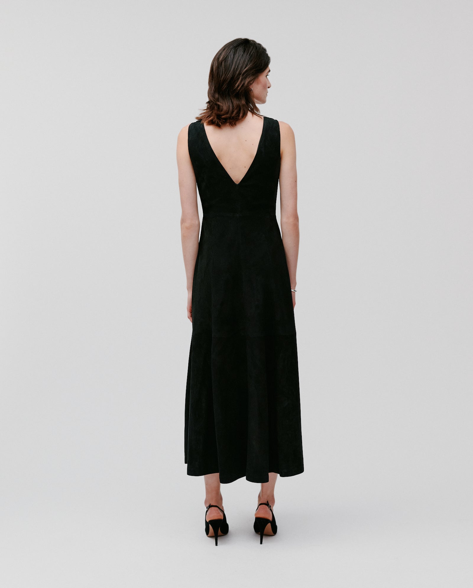 A woman stands facing away from the camera, wearing the IVY OAK LORENA DRESS—a sleeveless black dress featuring a deep V-back.