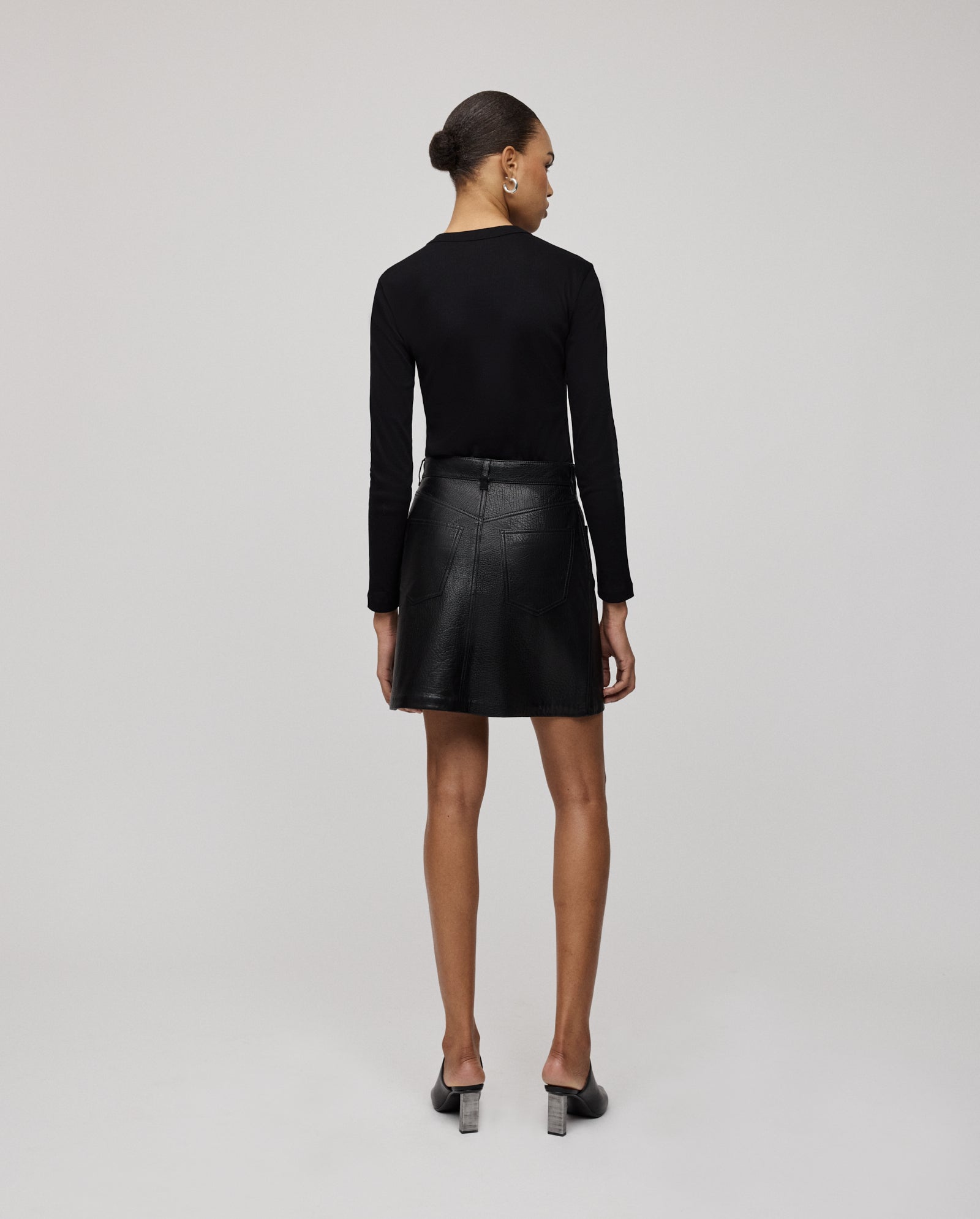 A woman stands with her back to the camera, wearing a black long-sleeve top and the IVY OAK LILOU Skirt, a premium hide A-line mini skirt.