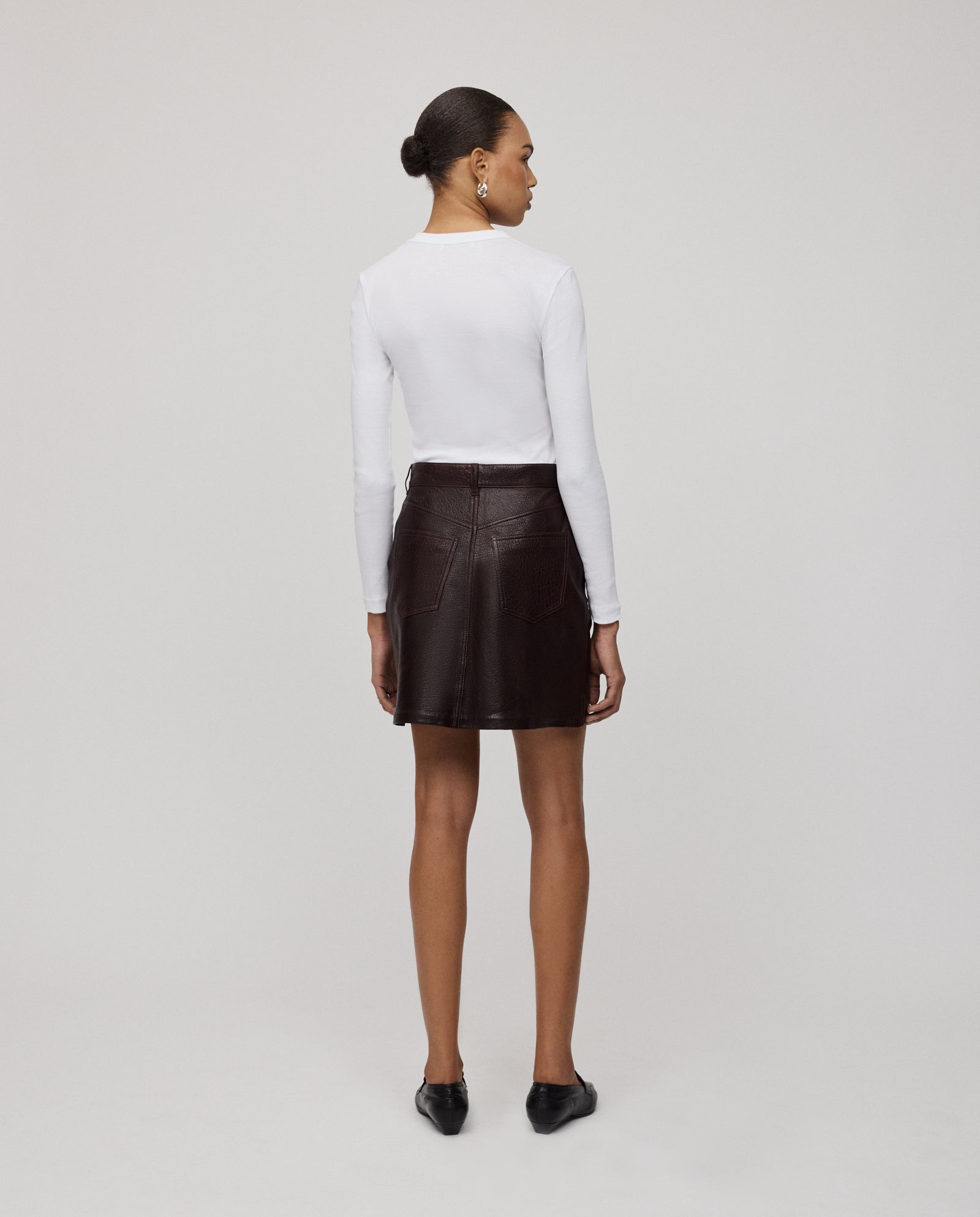 A woman stands facing away, wearing a white long-sleeve top, black shoes, and the IVY OAK LILOU Skirt in a dark brown A-line mini silhouette.